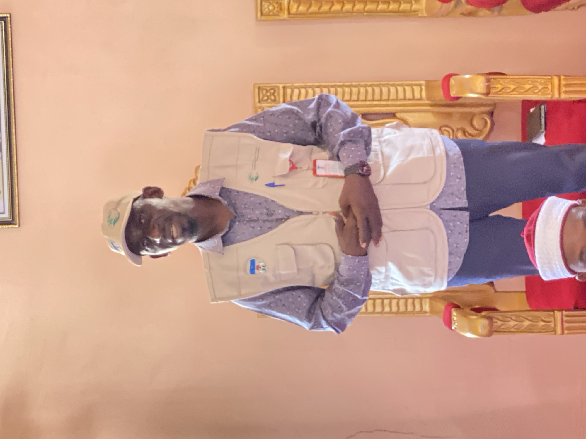 A middle-aged man is sitting on an ornate golden chair. He wears a light blue button-down shirt with a floral print, a beige vest, and a baseball cap. He is smiling and has his hands clasped in front of him. Another individual wearing a red and white cap is partially visible to the right. The background is a plain peach wall. The scene is likely indoors. The location is given as Sabon Tasha, Nigeria.