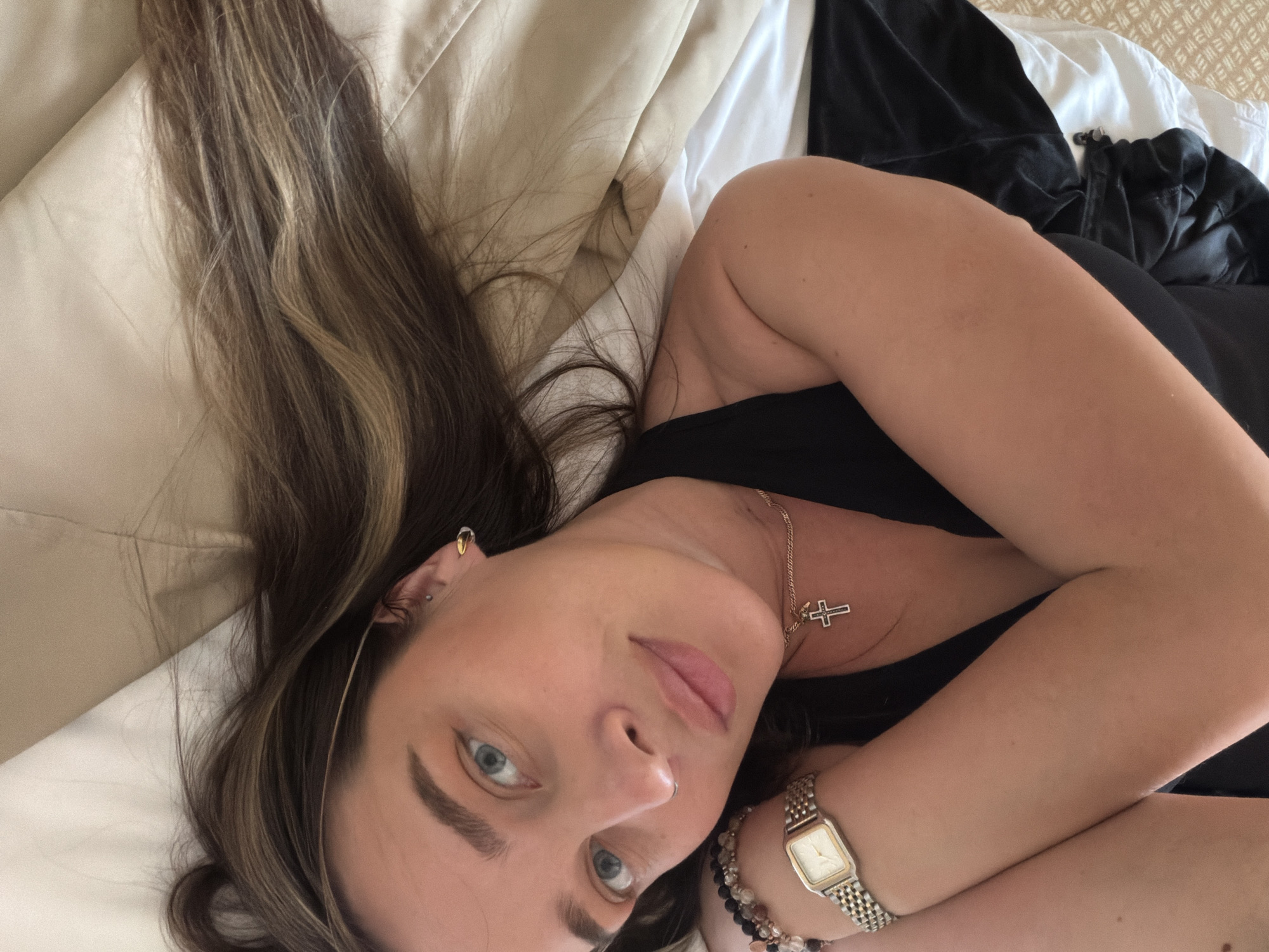 A woman is lying on a bed, viewed from an elevated angle. She has long, brown hair with highlights, fair skin, and light blue eyes. She is wearing a black top, a gold necklace with a cross pendant, multiple earrings in her left ear, and a watch and beaded bracelets on her left wrist. The bed has tan and white bedding. A black garment is visible on the bed near her torso. The location is Unknown, Unknown.