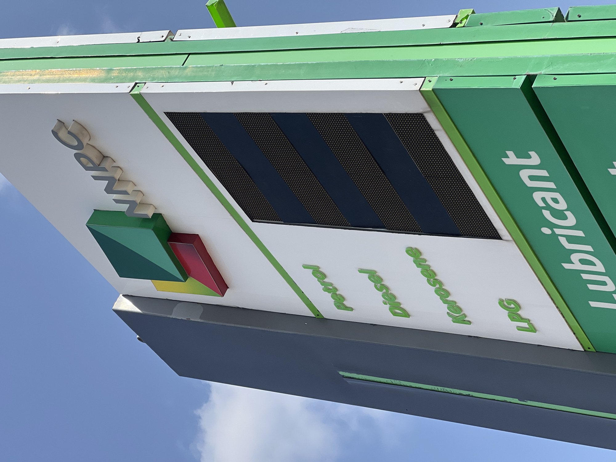 A low-angle, wide shot captures a NNPC petrol station sign against a clear blue sky with sparse clouds. The sign is large and predominantly white, featuring the NNPC logo in the upper right corner, composed of three colored geometric shapes: green, yellow, and red. Below the logo, the letters "NNPC" are raised in silver.
The main body of the sign lists fuel types vertically. "Petrol" is at the top, followed by "Diesel," "Kerosene," and "LPG." These words are rendered in a light green, three-dimensional font. To the right of these fuel names is a large, dark, digital display screen with a grid pattern, likely showing fuel prices. This screen is segmented by dark blue diagonal stripes.
The sign is supported by a vertical structure. The left side of the sign structure is a tall, dark gray panel. The right side is a bright green, painted metal frame with visible bolts. A green beam extends horizontally from the upper right corner of the sign structure.
At the bottom of the sign, a green panel displays the word "Lubricant" in large white letters.
The overall scene suggests a typical day at a Nigerian fuel station, given the presence of the NNPC branding, which is the Nigerian National Petroleum Corporation. While the specific location within Jalingo, Nigeria is not explicitly indicated on the sign itself, NNPC stations are prevalent throughout the country. No people are visible in the image. The environment is outdoor, under natural daylight.