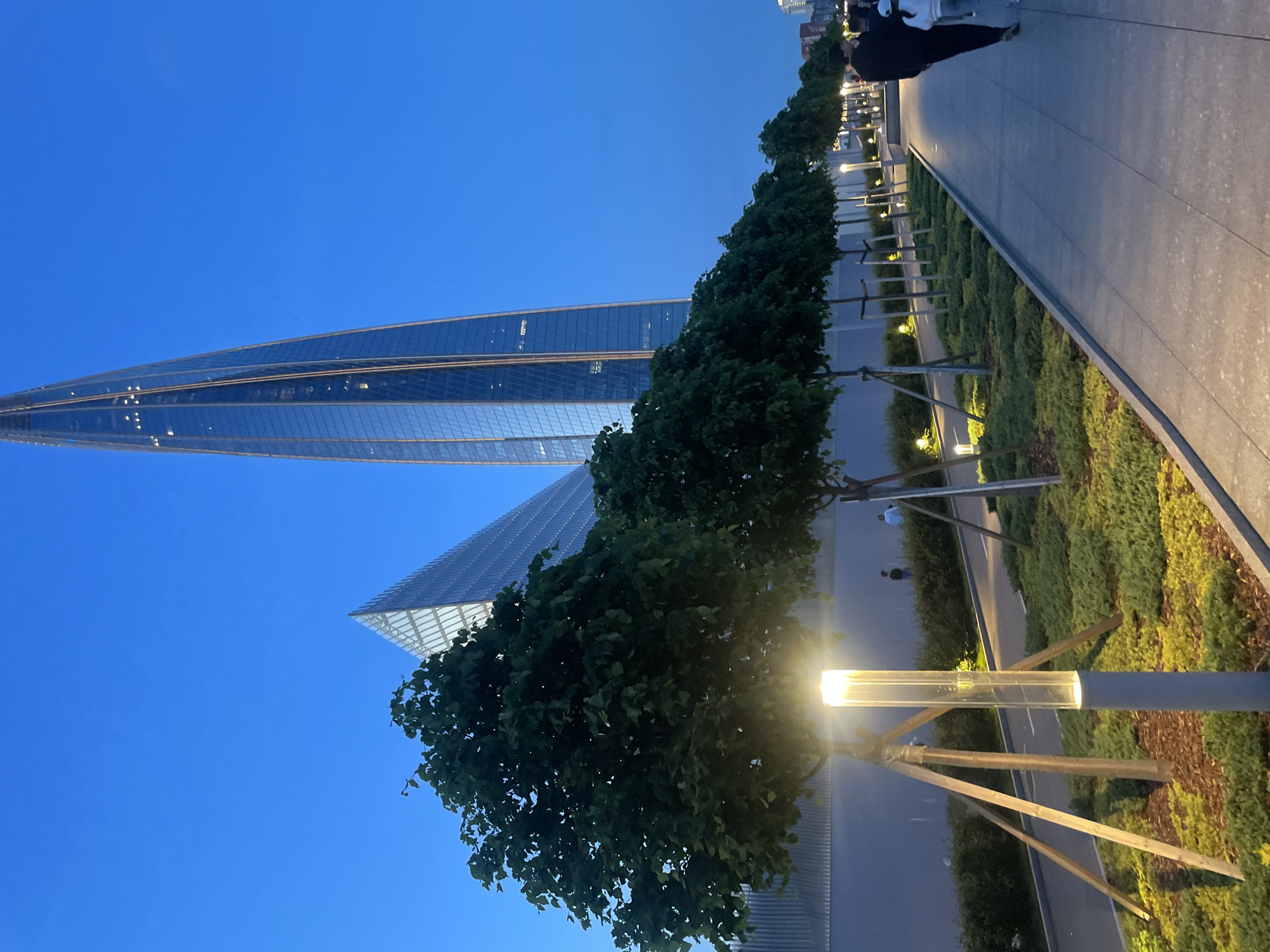 The image shows a view of the Lakhta Center skyscraper in Saint Petersburg, Russia, at dusk. The skyscraper is a modern, glass-covered building with a distinctive spiraling shape. In the foreground, there is a row of trees planted along a paved walkway. Modern streetlights illuminate the walkway and the surrounding landscaped area, which is covered with low-lying green plants and wood chips. In the background, a smaller triangular building, also clad in glass, is visible. A few people are walking along the path and in the distance. The sky is a deep blue, indicating twilight.