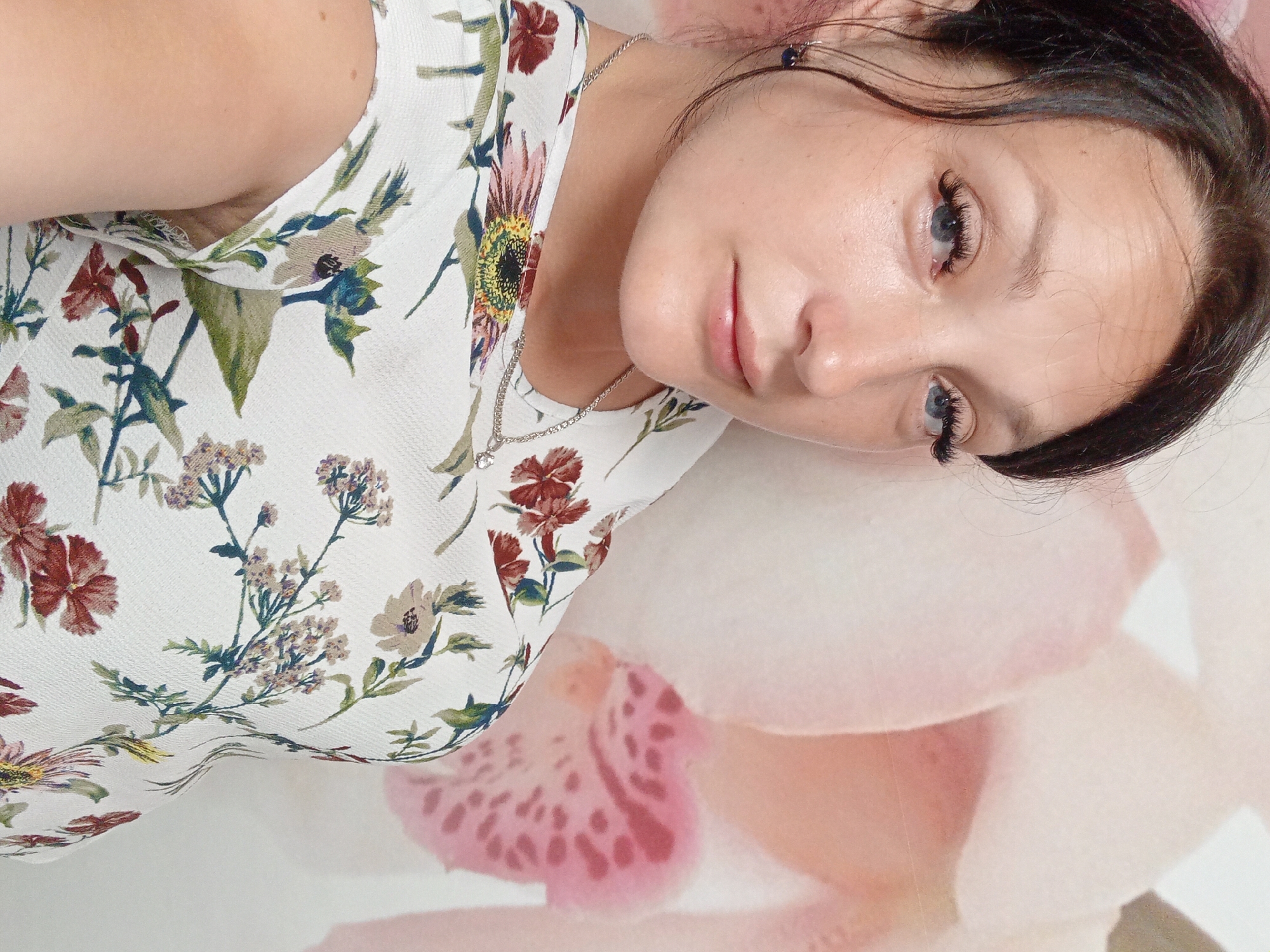 A medium shot shows a woman with dark hair and blue eyes from the shoulders up, looking to her right. She is wearing a white dress with a floral pattern and a silver necklace. The background is a light-colored wall with a large pink floral design. The image appears to be taken indoors. There is no discernible location in the city of Chuhuiv, Ukraine.