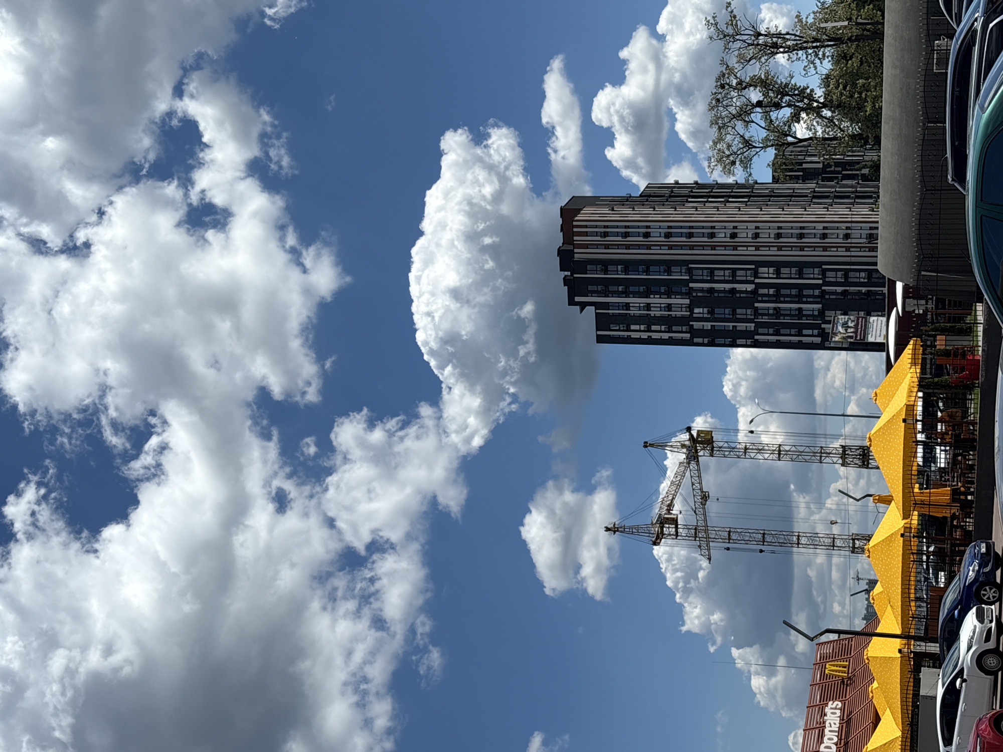The image, taken in daylight, showcases a portion of Hatne, Ukraine. Prominently visible are cumulus clouds filling the blue sky, partially obscuring a tall, modern apartment building with a black and white facade. Two construction cranes are positioned near the building, indicating ongoing development. In the foreground is a McDonald's restaurant with yellow umbrellas covering outdoor seating. Several parked cars are also visible.