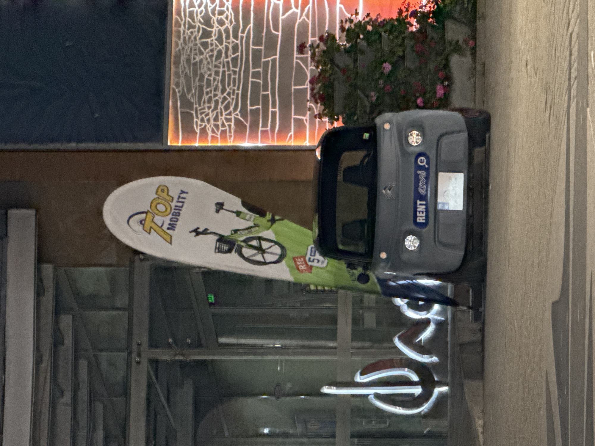 A grey electric microcar is parked in front of a building with glass doors and a sign. The car has a "RENT" sticker on its bumper. A tall, tear-drop shaped banner advertising "TOP MOBILITY" and featuring a green bicycle is leaning against the building next to the car. In the background, there is a wall decorated with a lit, crackled pattern and a planter filled with pink flowers. The scene is illuminated by artificial light, suggesting it is nighttime. The building appears to be in an urban setting, possibly a street in Burgas, Bulgaria, given the "rent.a.car" branding.