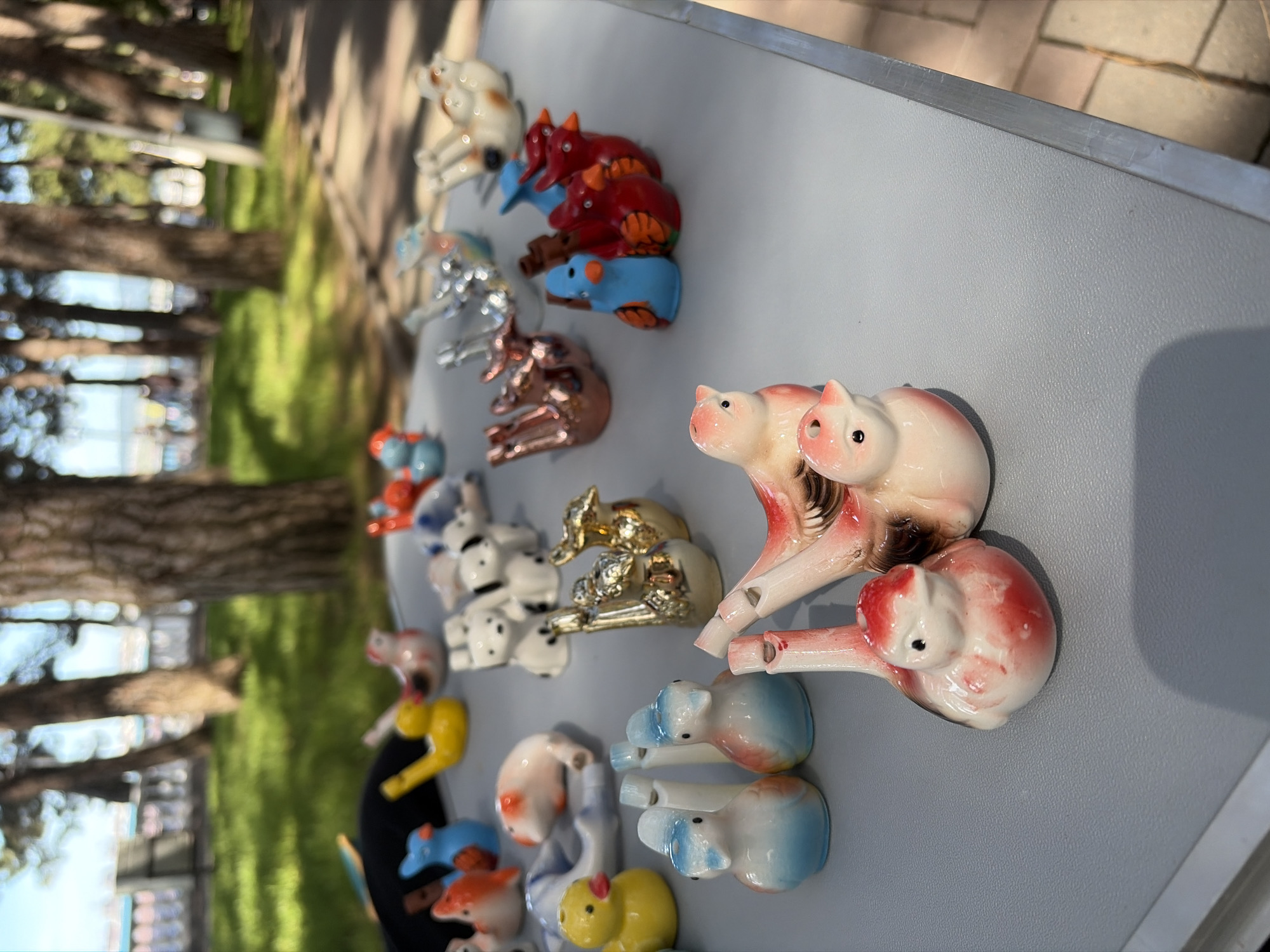 A close-up, eye-level, outdoor shot captures an assortment of ceramic bird figurines displayed on a light blue, textured table. The figurines are varied in color and design, with some depicting red and white birds, blue and white birds, and yellow ducks. In the background, blurred trees with green foliage and a hint of blue water suggest a park or waterfront setting in Gelendzhik, Russia. The ground appears to be paved with light-colored stones. The lighting indicates it is daytime, likely sunny, casting soft shadows.