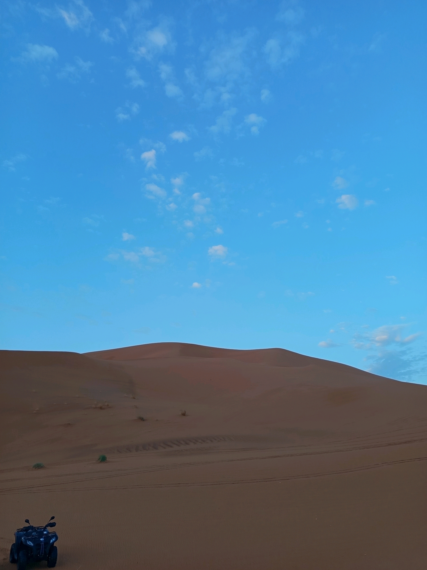 A desert landscape under a clear blue sky with scattered white clouds. In the foreground, a dark-colored ATV is parked on the sand. The middle ground features rolling sand dunes with tire tracks visible in the sand, indicating recent activity. The background is dominated by a large, smooth sand dune that extends towards the horizon. The scene suggests a recreational setting in a desert environment, likely near Rissani, Morocco, known for its desert terrain.