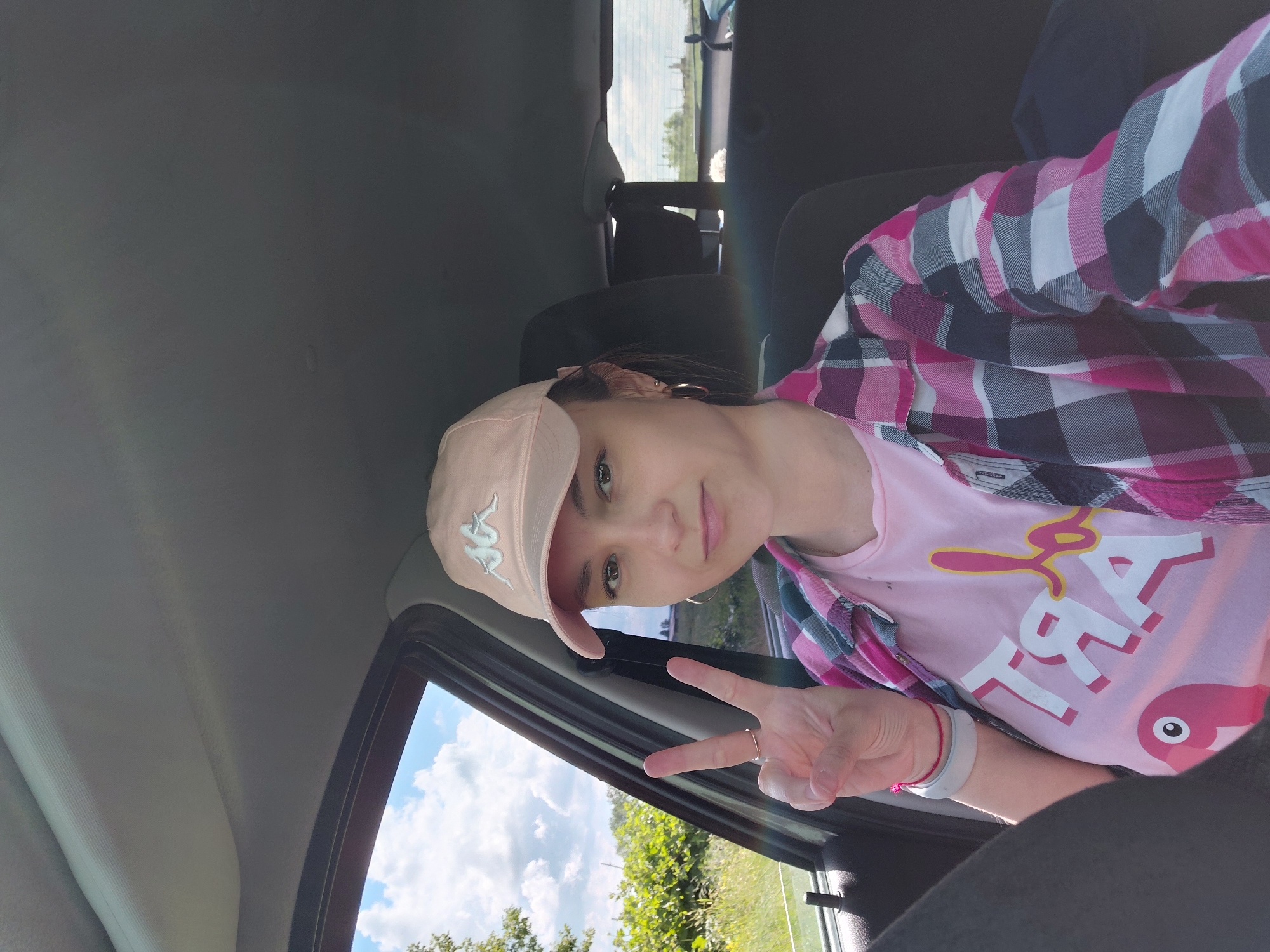 A young woman is sitting in the driver's seat of a car, looking at the camera and making a peace sign with her left hand. She is wearing a pink baseball cap, a pink t-shirt with the word "ART" and a flamingo graphic, and a pink and white plaid shirt. The car's interior is visible, including the ceiling, dashboard, and seats. Outside the car window, there is greenery and a cloudy sky. The sun is shining, creating a rainbow-like lens flare in the image. The scene appears to be in a rural or suburban area, possibly on a sunny day. The location of Tashkazgan, Russia is not discernible from the image.