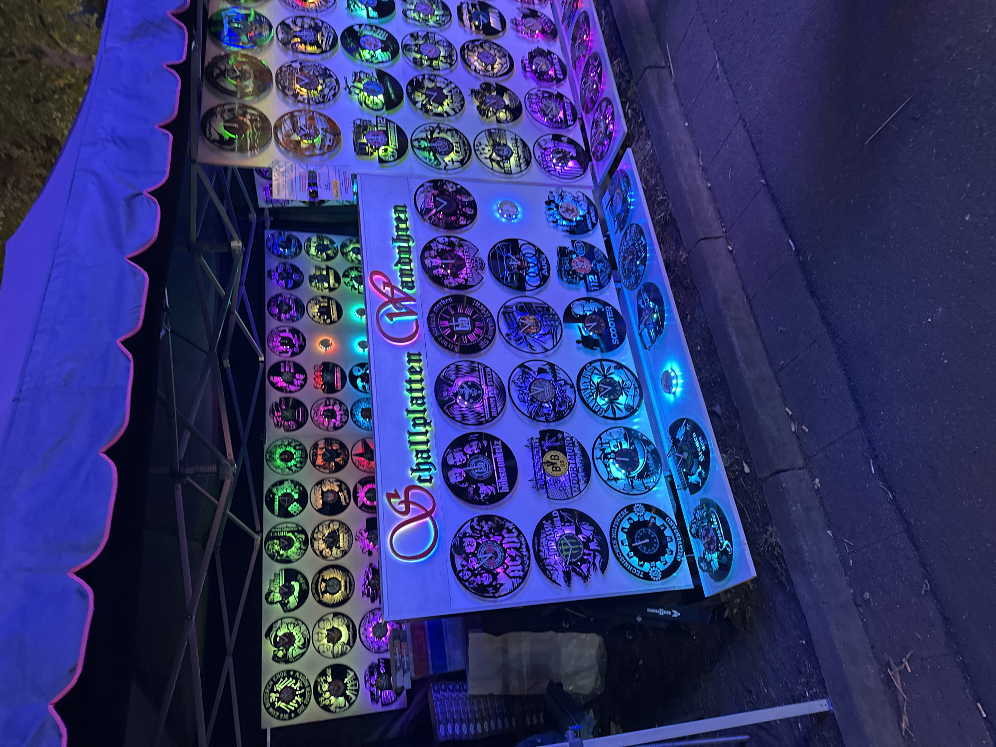 This image shows an outdoor stall at night in Bruchhausen-Vilsen, Germany, displaying a variety of illuminated decorative items, likely wall art or clocks, arranged on white shelving. The stall is covered by a blue tent with a pink scalloped trim. The items themselves are circular and feature a range of designs, including band logos (ACDC, Rammstein), movie themes (Jurassic World), and various other graphics and text. Many of these circular items are lit from behind with different colored lights, creating a vibrant and eye-catching display. The items appear to be made of metal and have intricate cut-out designs. The stall is set up on a pavement area next to a street, indicated by the curb and asphalt visible in the foreground. There are no people clearly visible in the image, only the displayed merchandise. The overall atmosphere is that of a night market or outdoor exhibition.