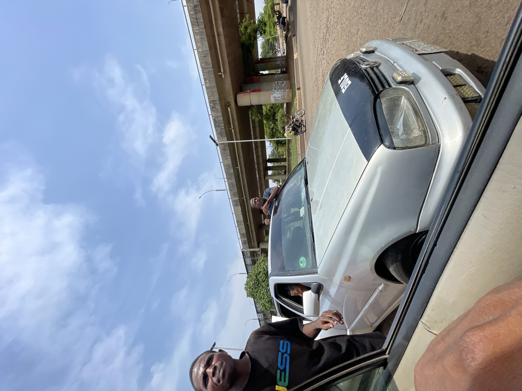 The image depicts a scene in Abuja, Nigeria, under a clear sky with scattered clouds. A two-lane elevated highway structure is visible. Two men are observed in the foreground near or inside a silver-colored car, with one leaning out of the window. The second man is on the car's roof. A woman sits inside the car, visible through the windshield. The person taking the picture is inside another vehicle and close to the car.