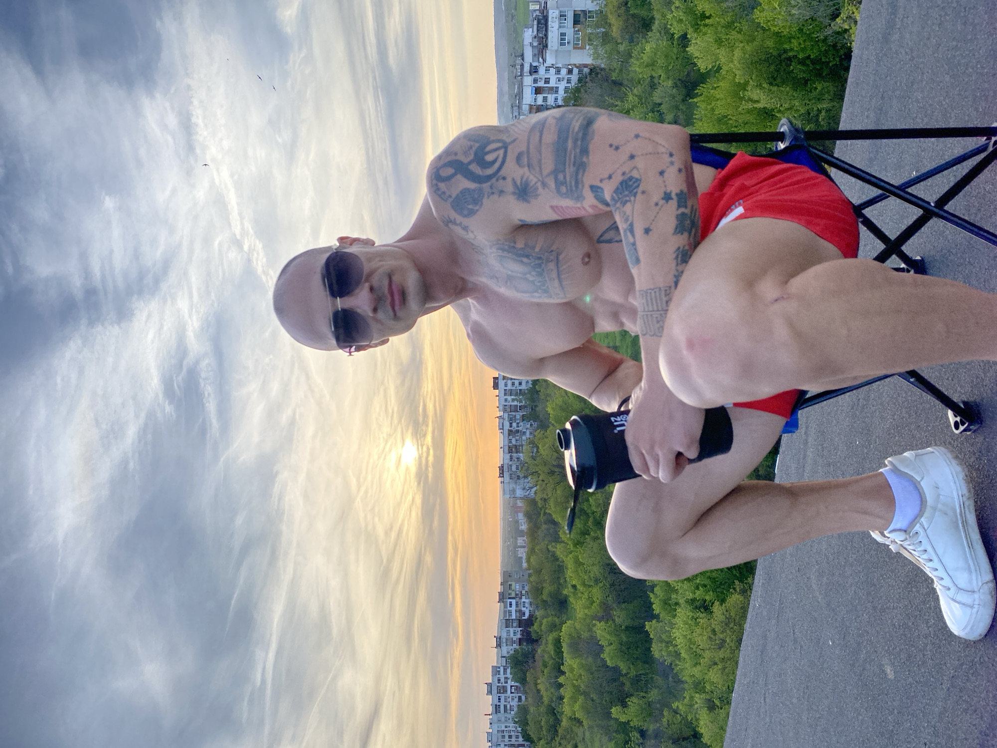A muscular, bald man with dark sunglasses is captured in a striking outdoor shot, sitting on a black folding chair on what appears to be a rooftop or elevated platform. He is bare-chested, revealing extensive tattoos covering his arms and torso, including partially visible text on his bicep. He wears red shorts and white sneakers with white socks, and holds a black water bottle with "NOON" visible on its side. He looks directly at the camera with a confident, neutral expression.
The scene unfolds during either sunrise or sunset, indicated by the low sun casting a golden glow across a partly cloudy sky. Delicate, wispy clouds stretch across the horizon, and a few birds can be seen in flight. Below the man, a sprawling urban landscape of apartment buildings is interspersed with dense green trees, suggesting a city park or forest area. The perspective offers a panoramic view, hinting at a location like Pleven, Bulgaria. The man's attire and the water bottle suggest he may be taking a break after physical activity.