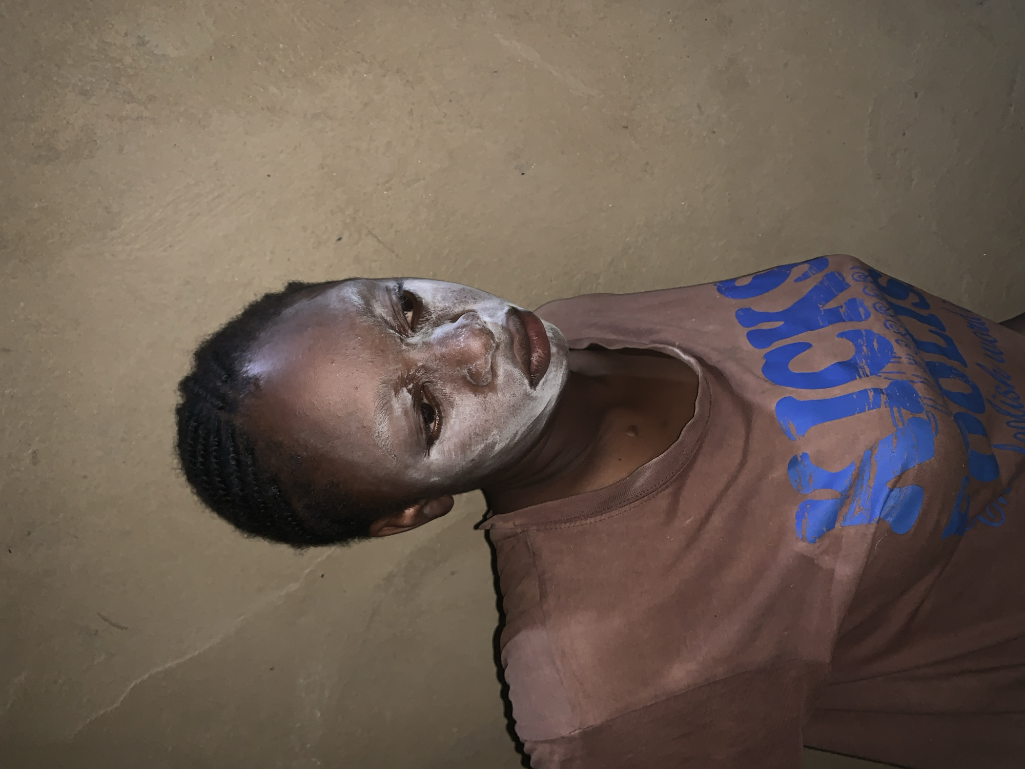 A person is lying on their side on a surface. The person has dark skin, braided hair, and a white substance smeared on their face. They are wearing a brown t-shirt with blue lettering. The background appears to be a wall or ground of a rough, textured material. The location is Agban, Nigeria.