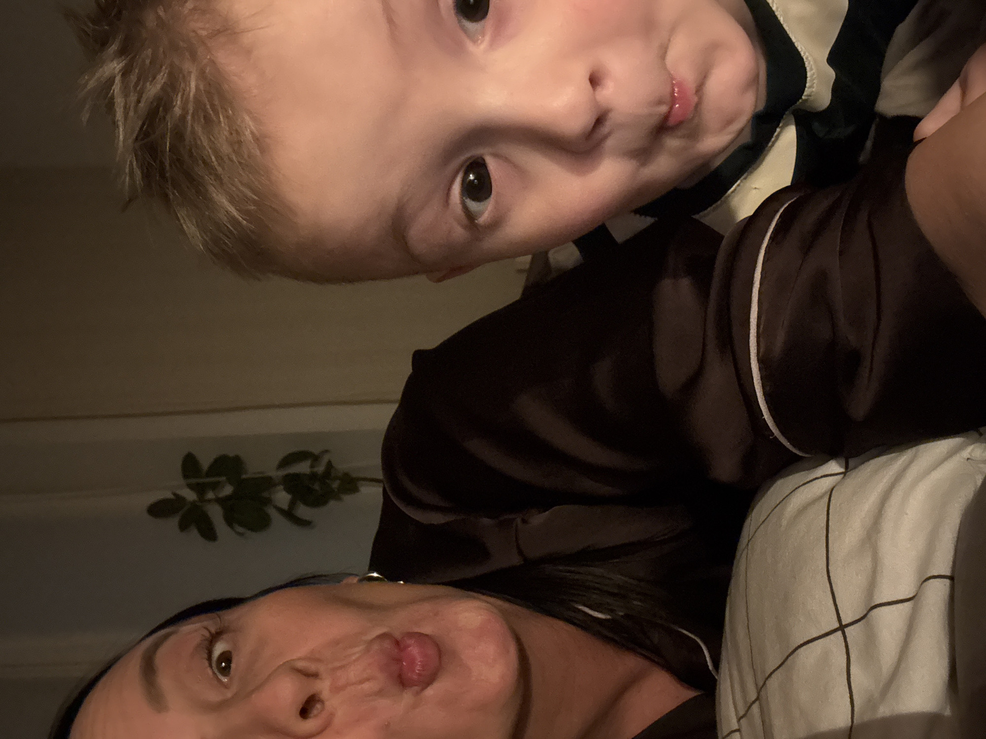 The image shows a selfie of a woman and a child, possibly in a domestic setting. The woman is at the bottom of the frame, and the child is positioned above her. Both are looking at the camera with pursed lips. The woman has dark hair and is wearing a dark-colored top with white piping. The child has light blonde hair and is wearing a striped shirt. A plant can be seen in the background. A pillow with a black grid pattern can be seen to the woman's right. The location is unknown; the city of Chaikovsky, Russia cannot be independently verified based on the image.