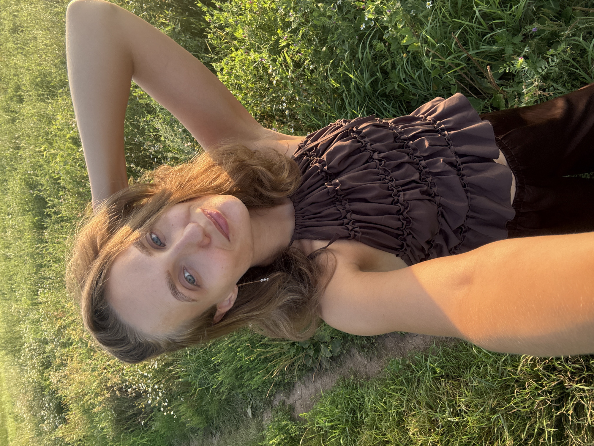 This is a vertical, medium shot selfie of a young woman lying on the grass in a field. The woman has blonde hair and light blue eyes. She is wearing a brown, ruched halter top and dark brown pants. Her left arm is bent and resting behind her head, while her right arm is extended, holding the camera. Her face is angled towards the camera, and she is smiling slightly. The background is a lush green field with some small white flowers scattered throughout. The lighting suggests it is late afternoon or early evening, with the sun casting a warm glow on the scene. There is no visible text in the image. The overall mood is relaxed and natural. The location context is Aksaitovo, Russia.