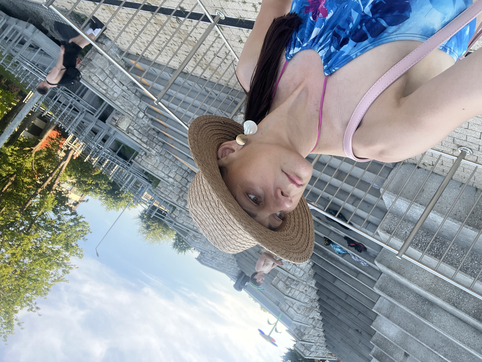 A woman wearing a blue floral swimsuit and a straw hat is posing for a photo. She has earrings in the shape of a white shell and a gold circle. A pink strap from her swimsuit is visible across her shoulder. The background shows a stone wall, metal railings, and concrete stairs. Trees and a bright blue sky with some clouds are also visible.
In the background, another person, a young boy, is visible sitting on the stairs, looking down. There are also some clothes scattered on the stairs. The image appears to be taken outdoors during the day. The lighting suggests it could be late afternoon or early evening. The overall scene seems to be a public area, possibly a park or a recreational space with steps and greenery.