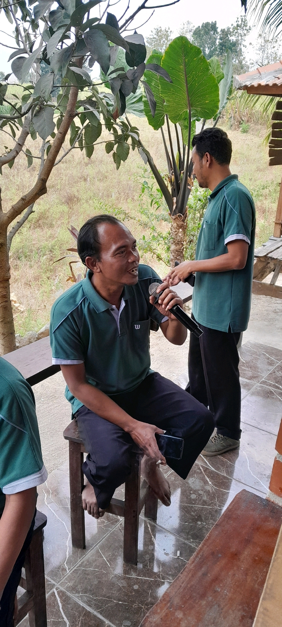 In Sidohasri, Indonesia, two men in green polo shirts are present in an outdoor setting. One man, seated on a wooden stool, holds a microphone and a smartphone, appearing to speak. He is wearing dark pants and is barefoot, with a cigarette in his hand. The other man stands nearby, facing the seated man and holding a smartphone. The environment includes trees, large green leaves, and a tiled floor.