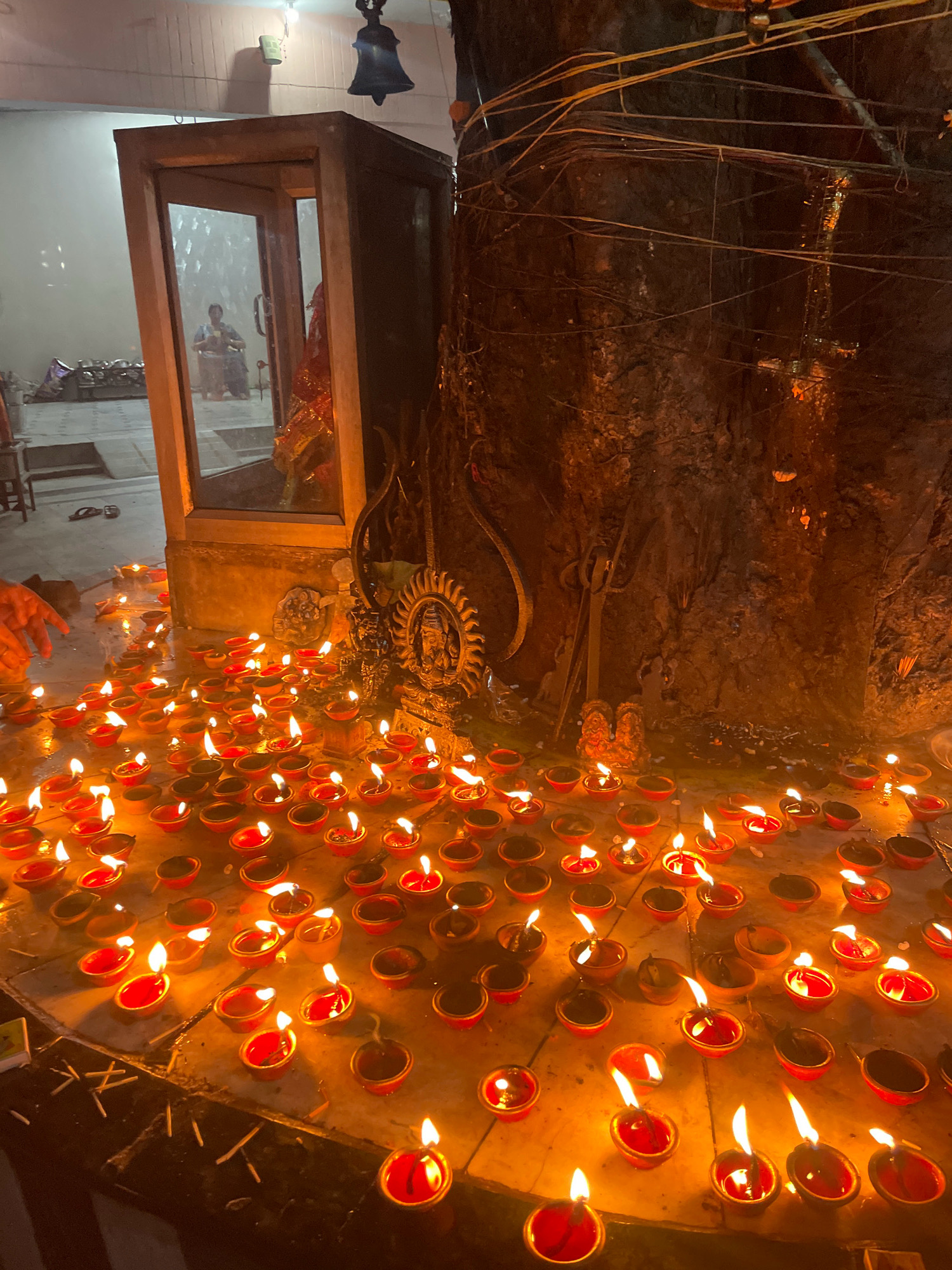 An indoor scene, likely a temple in Pathankot, India, is illuminated by numerous diyas, which are small clay oil lamps. The foreground is dominated by a large cluster of these lit diyas, arranged on a tiled surface. A large, textured tree trunk rises in the background, to the right of the frame, draped with wires and possibly other offerings. To the left, a glass-encased structure, possibly an altar or shrine, contains a seated figure, partially obscured. The overall ambiance is warm and spiritual, with the flickering flames of the diyas creating dancing shadows.