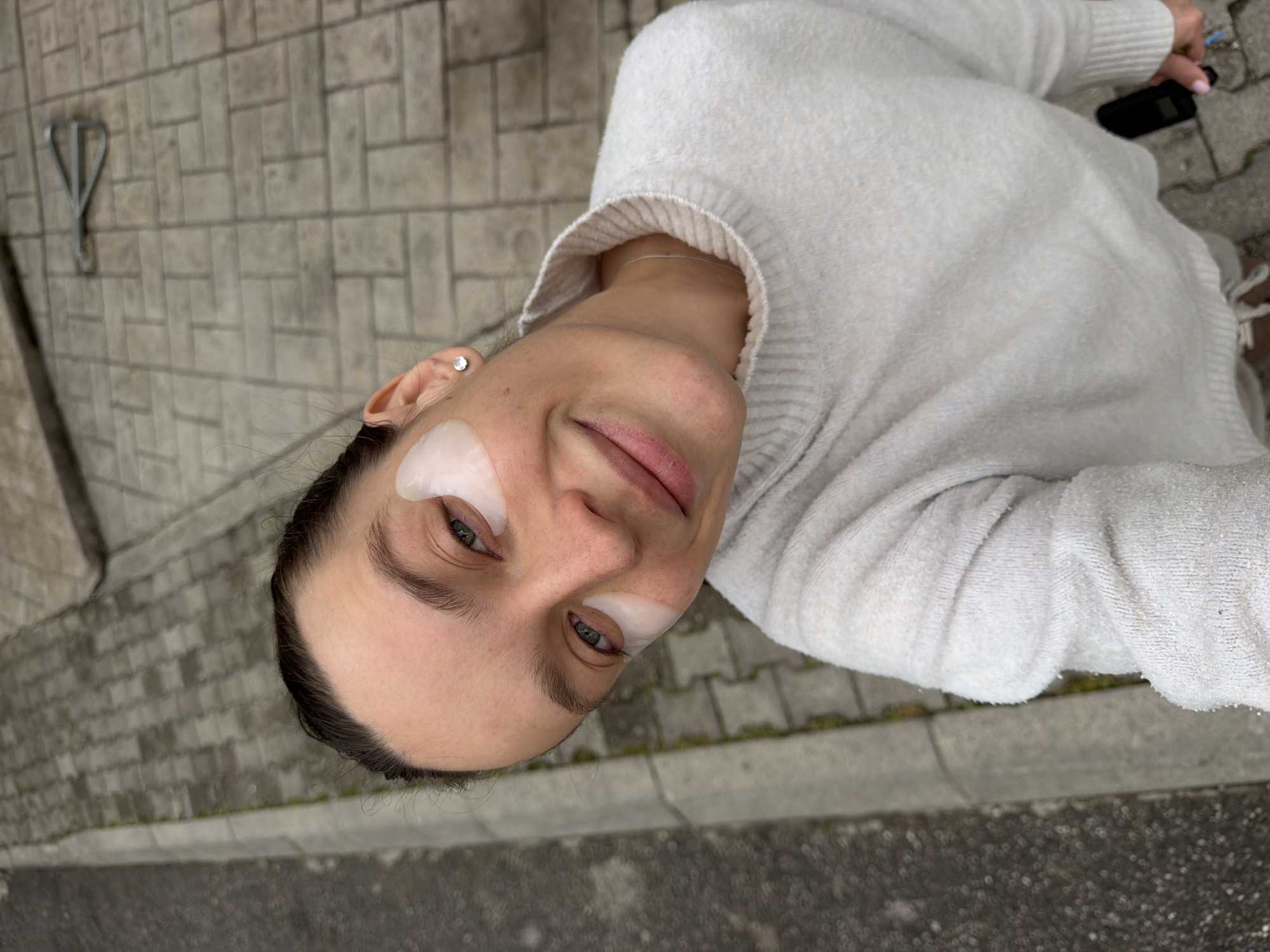 A woman is shown in an upward-facing selfie taken outdoors. She has fair skin and dark hair pulled back from her face. White, semi-transparent eye patches are affixed under both eyes. She is wearing a light-colored sweater with a ribbed collar. An earring is visible in her left ear. A black object, possibly a phone or electronic device, is held in her right hand. The background consists of gray concrete pavement and a textured wall with a metal bracket. The location is likely Budva, Montenegro based on visual features.