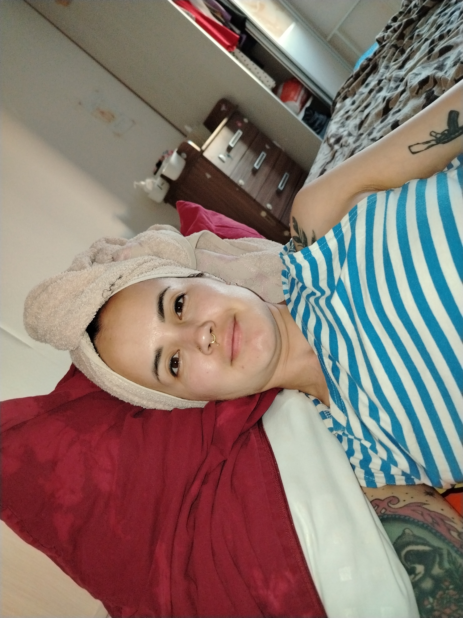 A woman is lying on a bed with a red pillow under her head and a towel wrapped around her head. She is wearing a blue and white striped shirt. Her left arm is visible, and she has a tattoo on it. There is a chest of drawers and a closet in the background. There is no information in the image to indicate that this is located in Noviy, Russia.