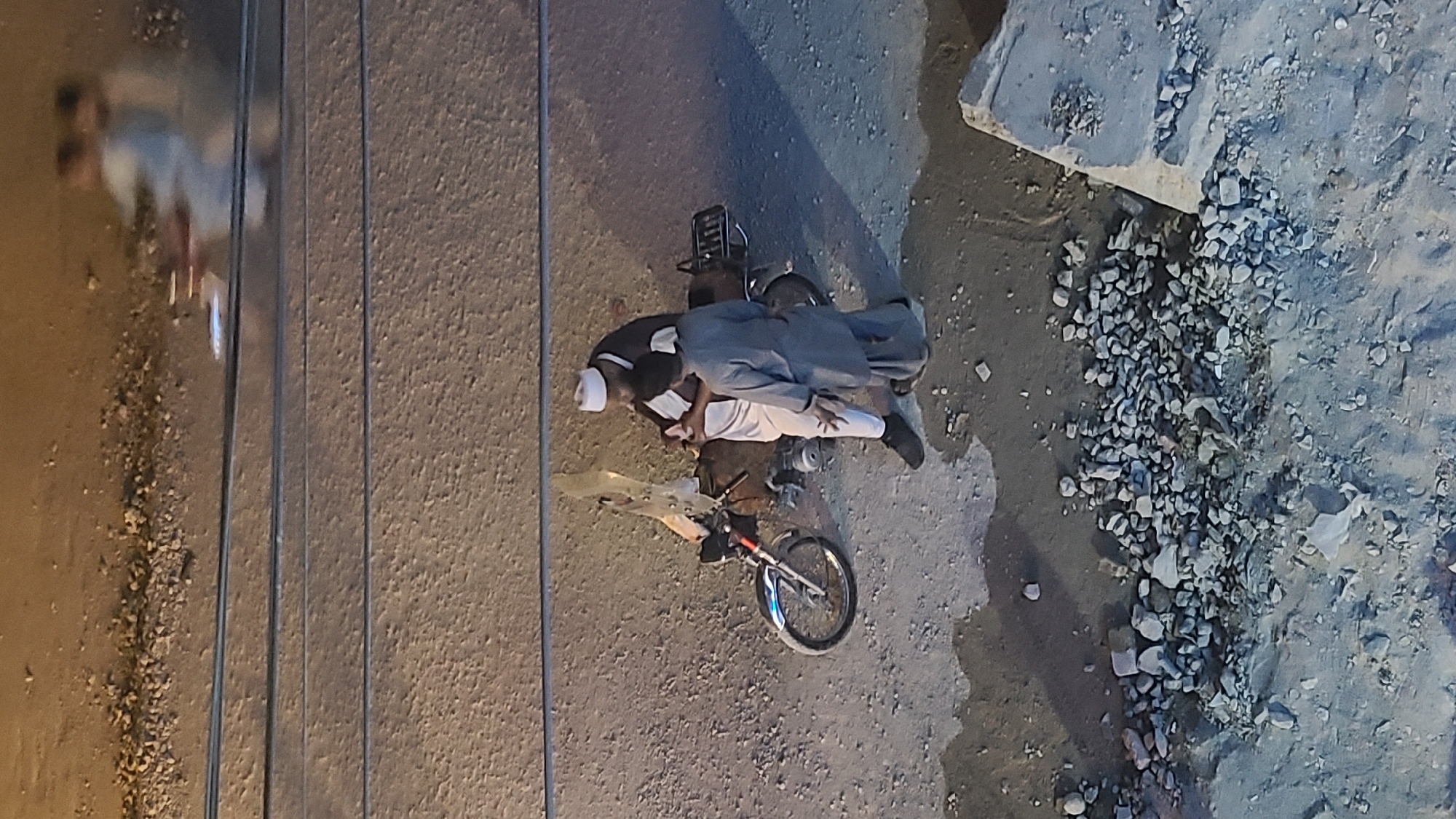 The image shows a scene from an elevated perspective, possibly taken from a building overlooking a street. In the center, two people are interacting beside a motorcycle. One individual wears a white head covering (possibly a taqiyah) and light-colored clothing, while the other is dressed in darker attire, possibly a shalwar kameez. They appear to be assisting each other near the motorcycle. The motorcycle is positioned on the edge of the street, with the foreground showing a rough patch of ground with scattered rocks and debris. Part of a bicycle with blue accents is also visible in front of the motorcycle. Along the left edge of the frame, electrical cables are seen running parallel to the street. The overall environment suggests an urban or peri-urban setting. Based on the query context, the location is within Quetta City Tehsil, Pakistan.