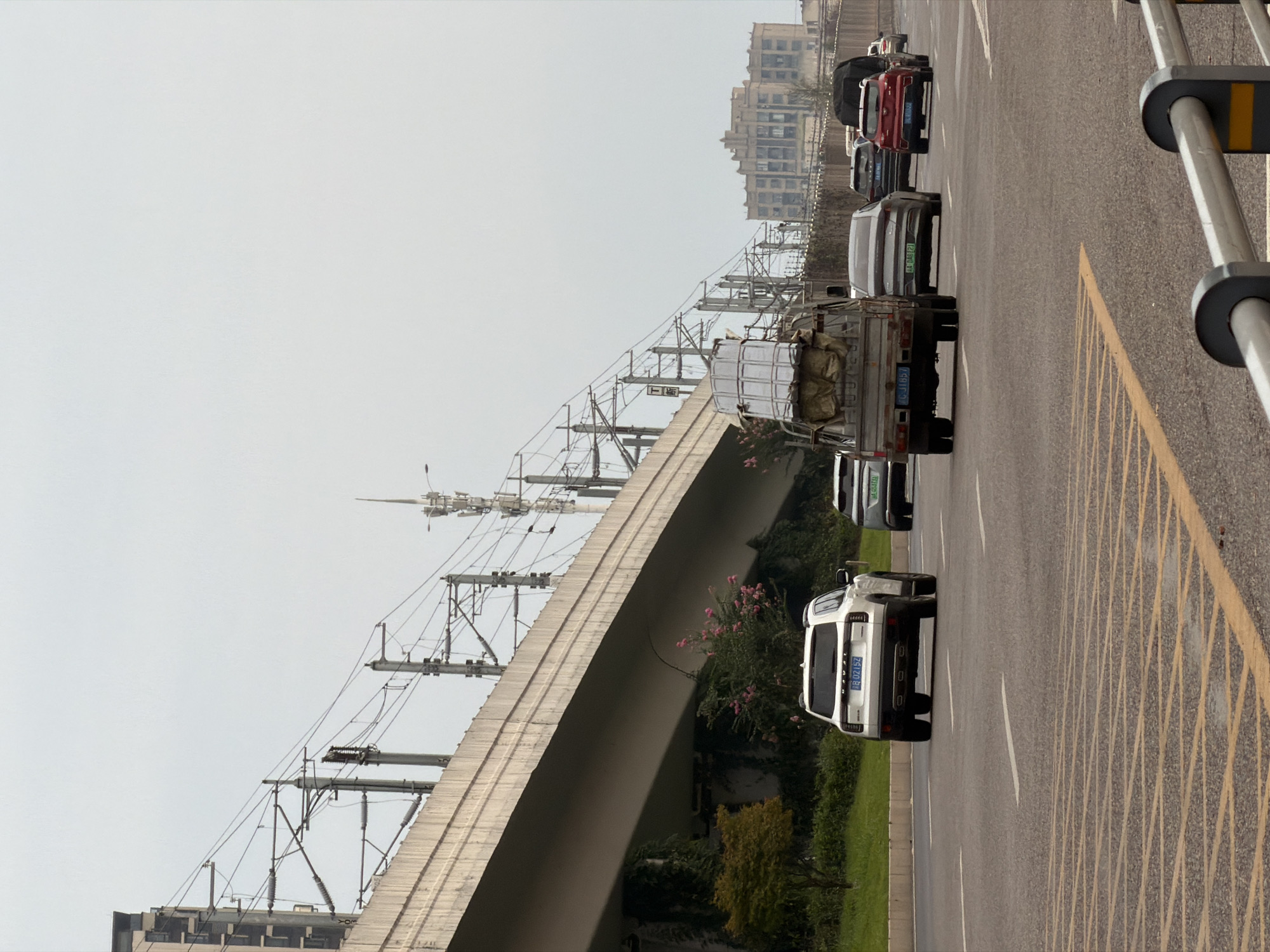The image depicts a street scene in Jiangjin District, Chongqing, China, on a cloudy day. A multi-lane road with yellow and white road markings occupies the foreground. Several vehicles are traveling on the road, including a white SUV with license plate "B 0Z152", a silver sedan, a red SUV, and a small truck carrying a load of what appears to be metal sheeting covered with tarpaulin. In the background, a large elevated concrete structure, likely an overpass or viaduct, runs diagonally across the frame, with numerous electrical wires and poles for a railway system suspended above it. Adjacent to the overpass are trees and vegetation, with some green ivy climbing the structure. Further in the distance, several buildings are visible, including a multi-story building with a distinctive architectural style. The sky is overcast, suggesting subdued lighting conditions.