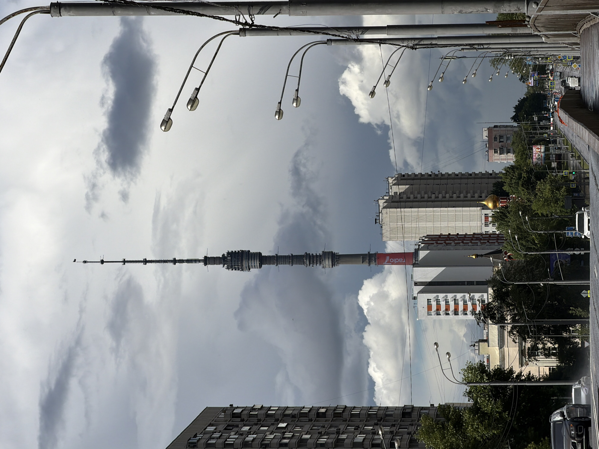 This is a vertical, eye-level, medium shot of a city skyline on a cloudy day. The foreground features a row of streetlights that extend upwards and out of frame. Beyond the lights, a large, imposing television tower dominates the center of the image, its complex structure reaching towards the sky. To the left and right of the tower are various buildings, ranging from modern high-rises to older apartment blocks. Trees and greenery are visible between some of the buildings, suggesting a mix of urban and park-like environments. In the background, a street scene with cars and traffic lights can be seen, indicating an active urban setting. The sky is a dramatic mix of dark, heavy clouds and patches of brighter sky, suggesting the weather is changeable. The overall impression is one of a sprawling metropolis under a vast, cloudy sky. The word "radio" is visible in red lettering on the side of the television tower. The image appears to have been taken in Moscow, Russia.