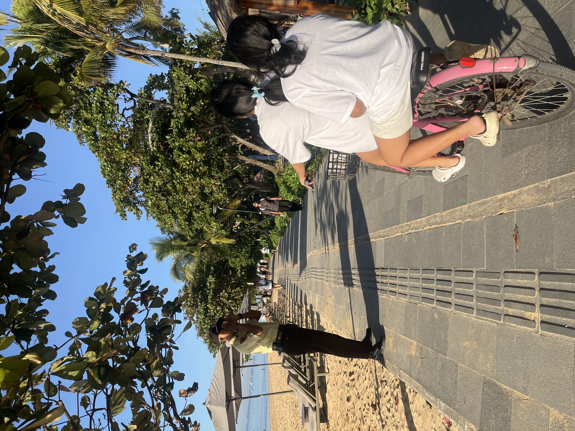 In Denpasar, Indonesia, a uniformed security guard stands on a sandy beach path, looking through a camera. He is wearing a light yellow shirt, dark pants, and a cap, with his hands cupped around his eyes as if to shield them from the sun or focus his vision. Beside him, a paved walkway runs parallel to the beach, lined with lounge chairs and umbrellas. Further down the walkway, two young women on a pink bicycle, wearing white t-shirts and shorts, are moving away from the camera. The bicyclist in front has a blue hair tie, and the one behind has a grey one. In the background, lush green trees and palm trees border the path, with a clear blue sky overhead. The scene suggests a sunny day at a popular beach resort area.