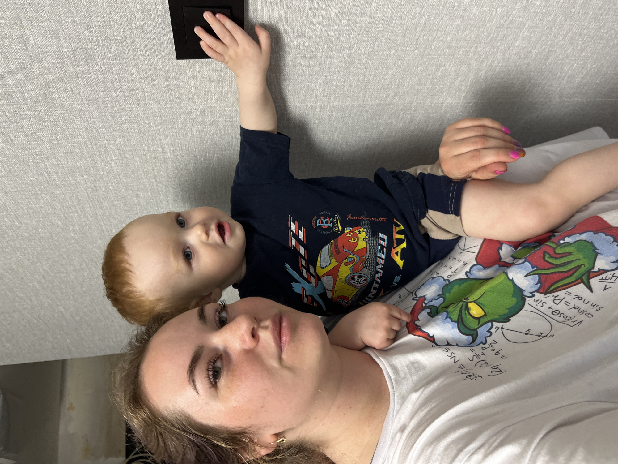 The image shows a woman and a young child lying down. The child is reaching for a light switch on the wall. The woman is wearing a white shirt and the child is wearing a navy blue shirt. The woman is lying on a white t-shirt with a green cartoon character on it. The location is not identifiable as the city of Chaikovsky, Russia, from the image.