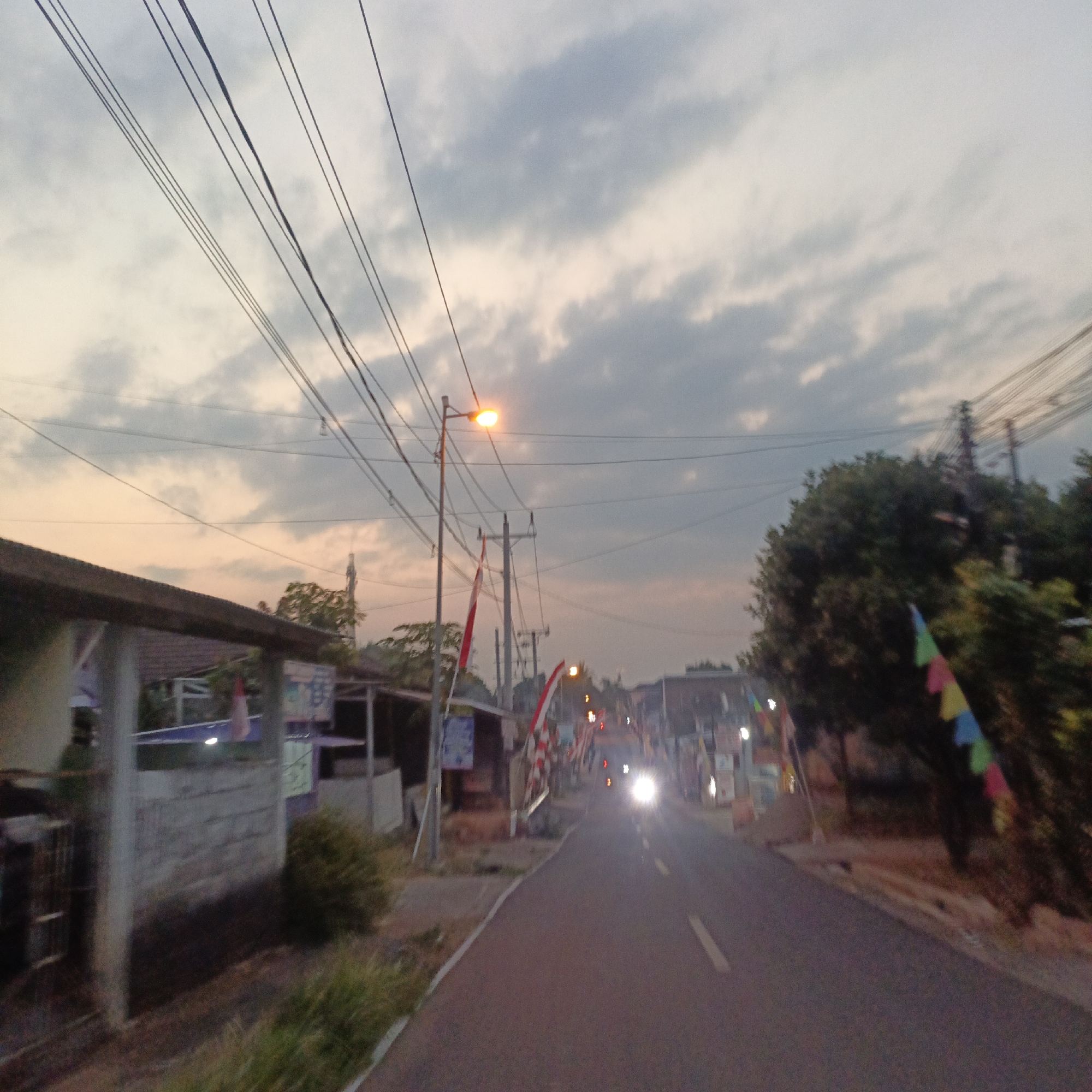 The image depicts a street scene in Salatiga, Indonesia, likely at dusk or dawn, given the soft, warm lighting of the sky and the illuminated street lamp. Overhead, numerous power lines stretch across the frame, casting shadows. The road itself is paved and appears to be a two-lane street.
On the left side of the road, there's a low-lying building with a dark, angled roof, possibly a shop or residence, constructed with grey concrete walls and some visible white pillars. Further down the left side, a small kiosk or stall is present, adorned with red and white flags. A street lamp with a bright orange light illuminates the area.
On the right side of the road, there are more buildings and dense foliage, including several trees. Colorful triangular flags are strung along the trees. A car is visible on the road, its headlights brightly illuminated, suggesting it's moving towards the viewer. Other lights are visible in the distance, indicating further structures or activity along the street. The overall impression is that of a residential or commercial area experiencing the transition from day to night or vice versa.