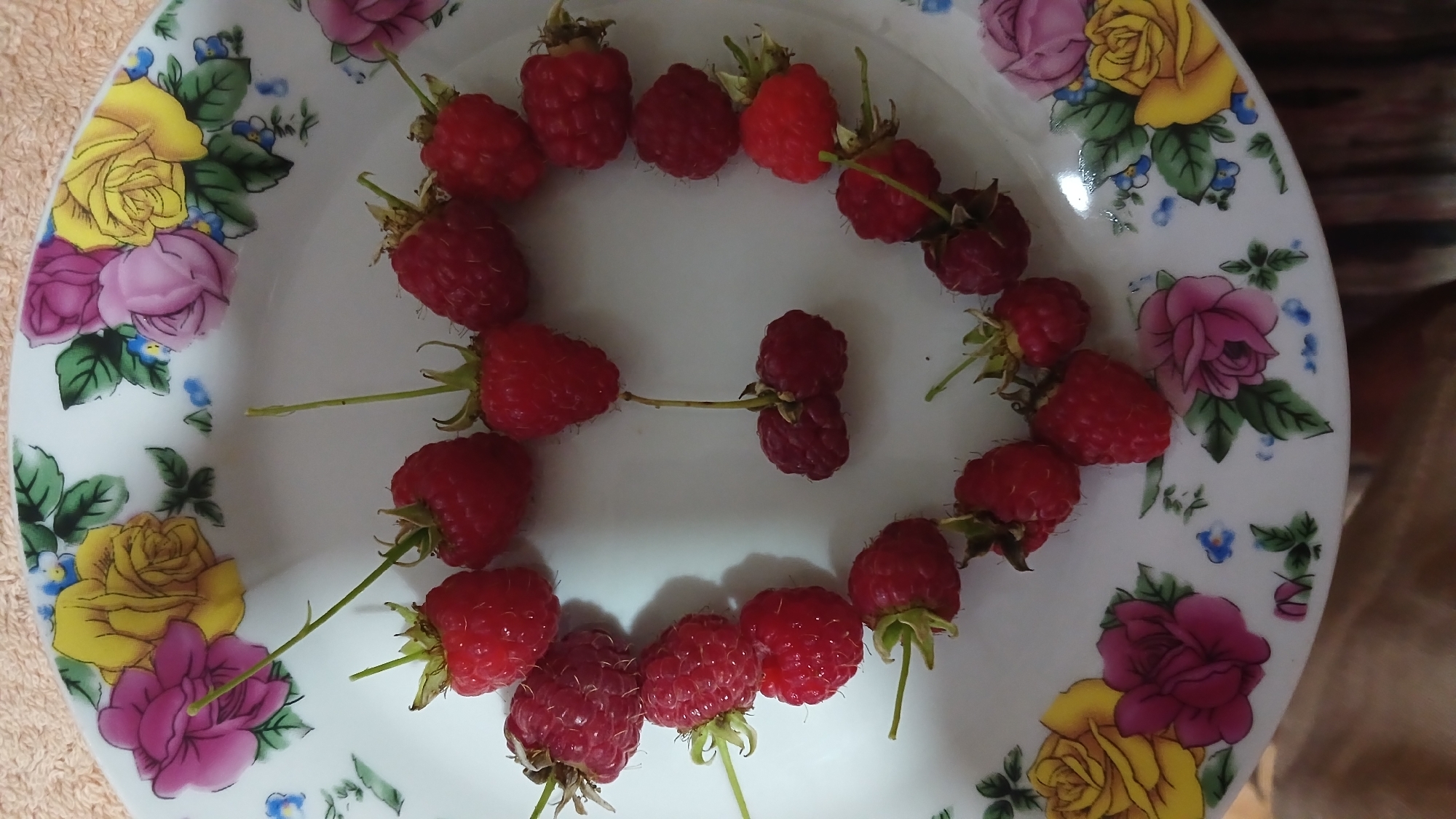 A close-up overhead shot shows a heart-shaped arrangement of raspberries on a white plate with a floral pattern. The plate is decorated with pink, yellow, and purple roses, as well as blue and green accents. The raspberries are red and appear to be fresh. The arrangement is centered on the plate, with some raspberries forming the outer edge and others filling in the shape. The lighting is natural, casting a soft shadow on the plate. The background is out of focus but appears to be a textured surface, possibly a tablecloth or countertop. The image was taken indoors in Monastyryska, Ukraine.