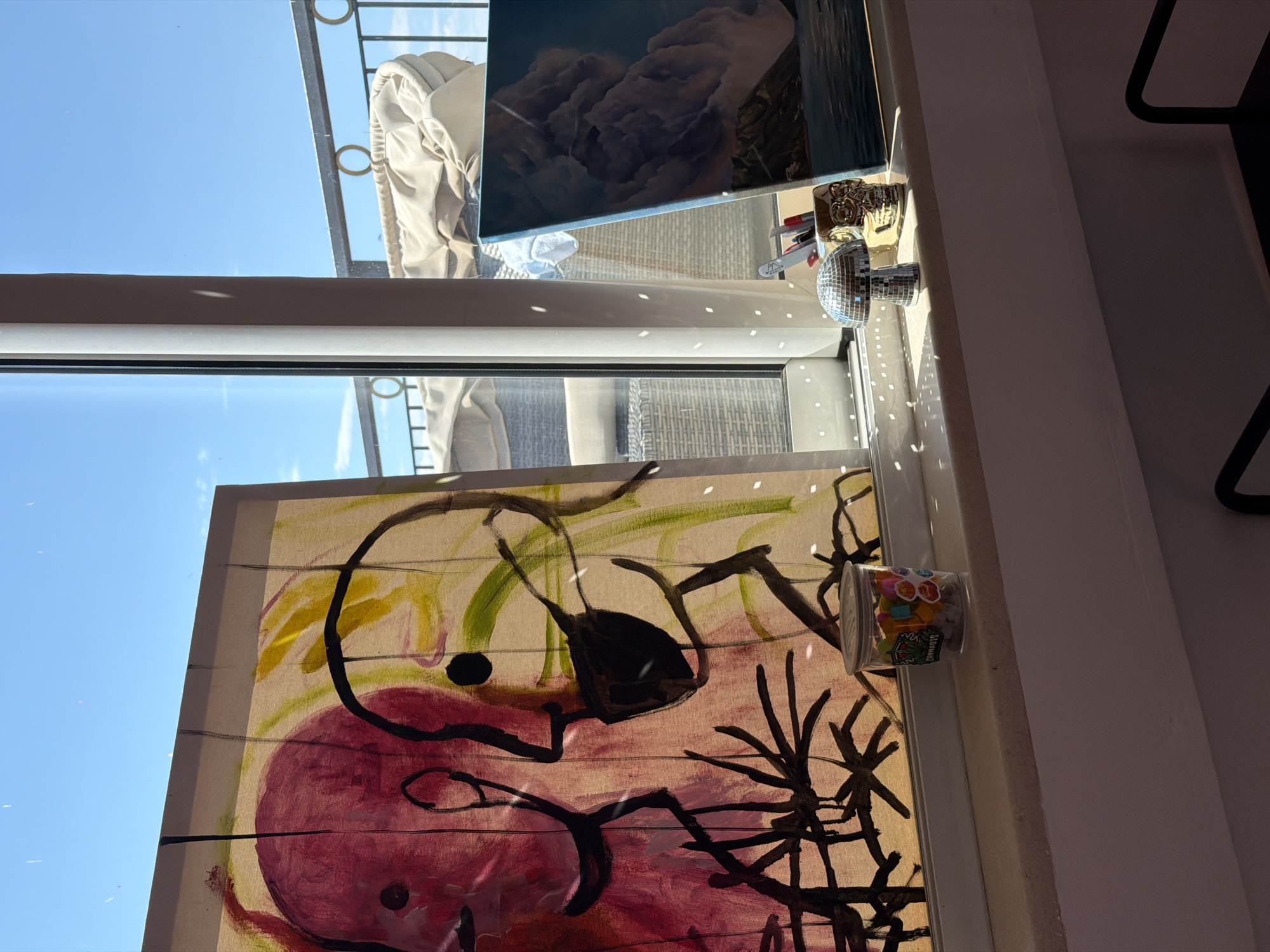 The image shows a view from an apartment window in Budva, Montenegro. The window sill contains several objects, including a disco ball, a painting depicting abstract figures in red, yellow, and black on a light-brown background, and a clear plastic container filled with colorful objects. Outside the window, a balcony with a metal railing is visible, as well as a building, a painting, and a clear blue sky. The window is reflecting the sky and objects in the room. The light is bright and creates speckled patterns on the sill from the disco ball.