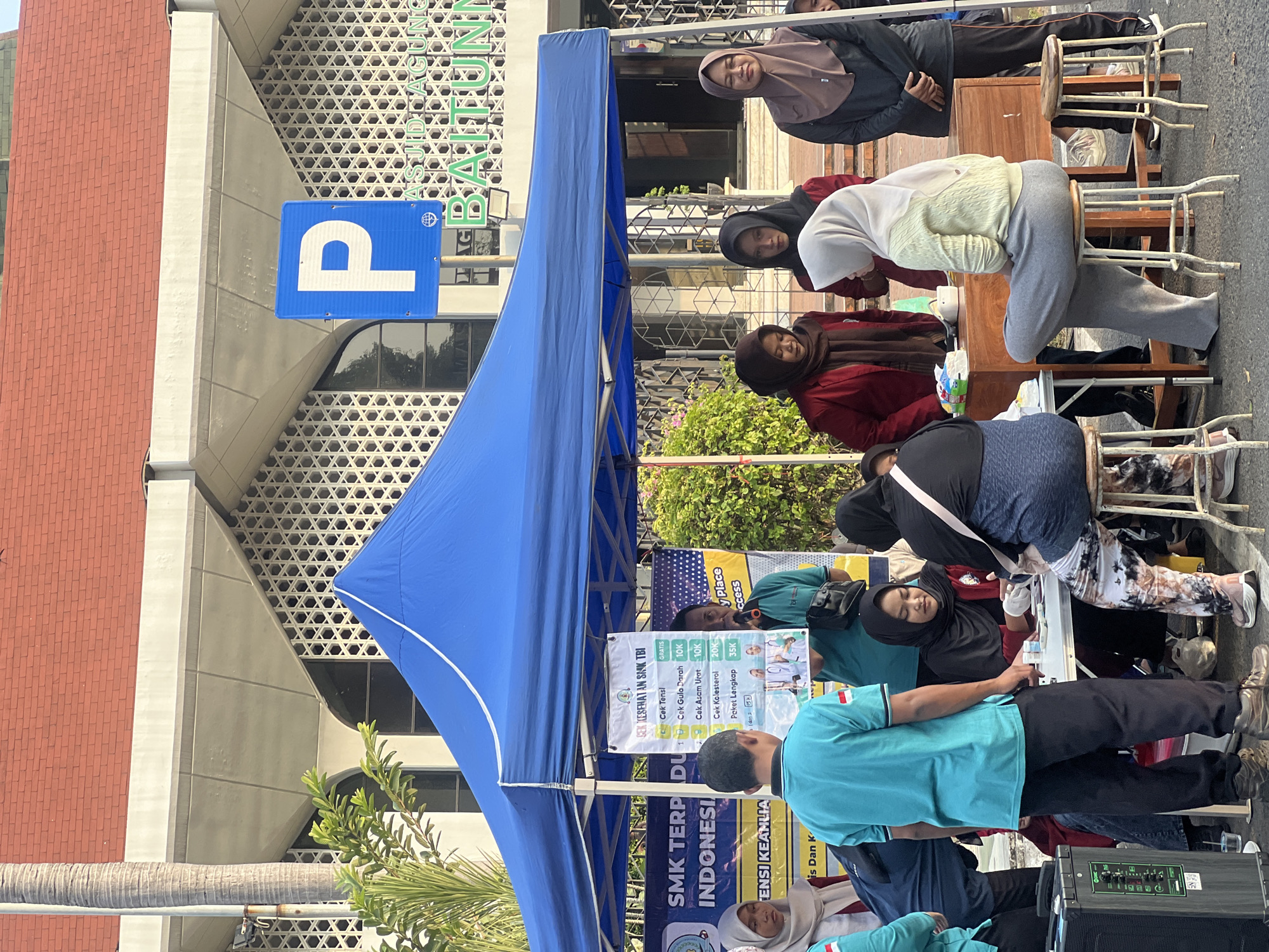 The image depicts an outdoor health screening event held in front of a building with a red tiled roof and a distinctive patterned facade, possibly a public institution or community center, in Kecamatan Pati, Indonesia. A large blue canopy tent is set up, providing shade for a table where several individuals are engaged in activities related to health checks.
At the table, a person wearing a black hijab and a red jacket is attending to a patient, indicated by the presence of medical supplies on the table, including what appears to be a blood pressure monitor and vials. Another person, seated on a stool and wearing a light-colored hijab and a yellow knitted sweater, seems to be receiving a service. Other individuals, some in hijabs, are standing around the table, observing or waiting their turn. One man, wearing a teal t-shirt and black pants, stands near the table, holding a sign with health check services and prices listed in Indonesian. Behind the tent, a sign displaying the letter "P" in a blue square, indicating parking, is visible. The background reveals the architectural style of the building and some greenery. The overall scene suggests a community health outreach program or a similar event offering accessible health services.