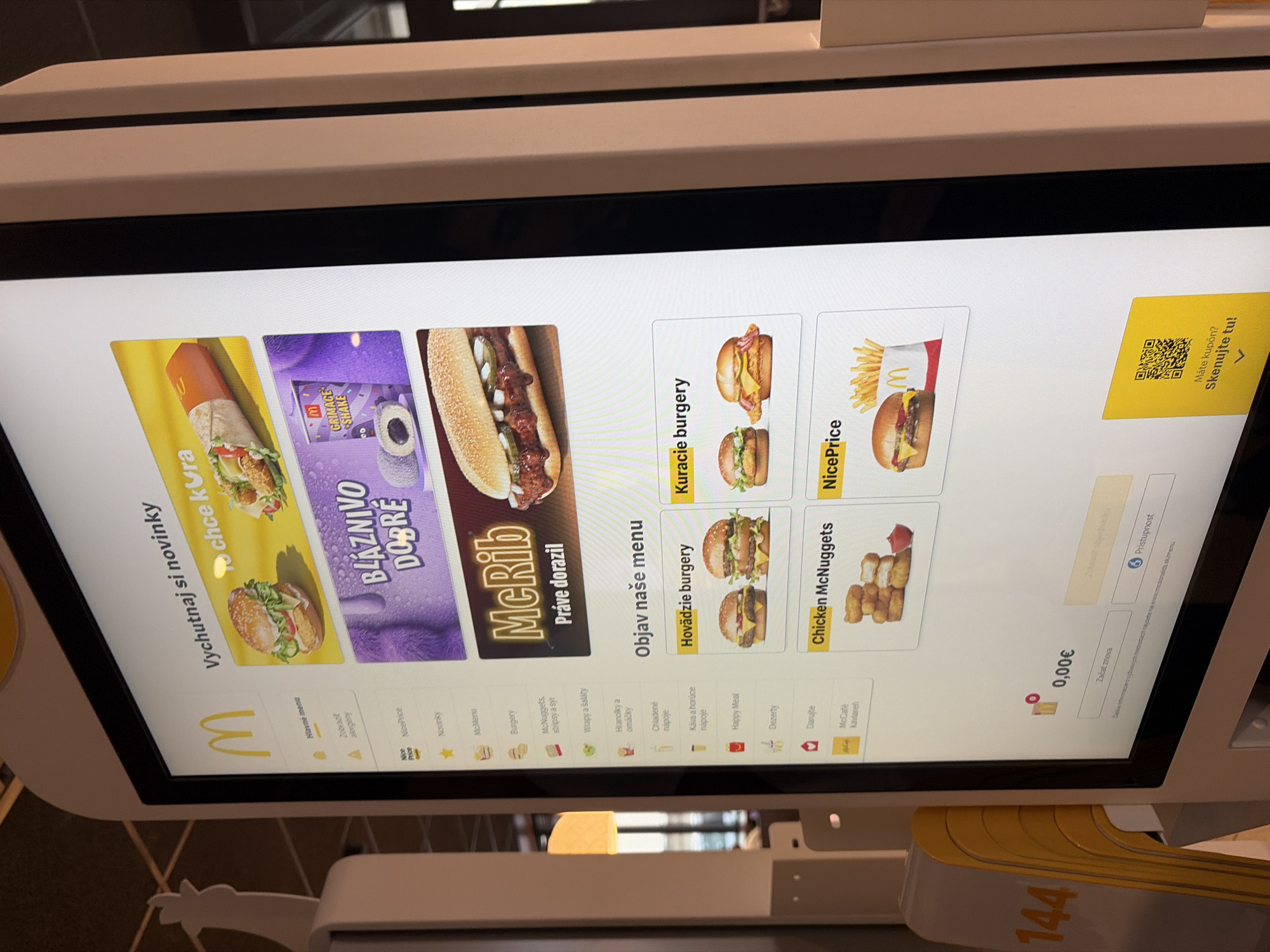 The image shows a close-up of a McDonald's self-order kiosk screen in Nitra, Slovakia. The screen displays various menu items, including burgers, chicken nuggets, and drinks. There are promotions for new items like "Bláznivo dobré" (Crazy good) and "McRib." The kiosk is located indoors, likely within a McDonald's restaurant. The screen is bright and colorful, with images of food and promotional graphics. The overall scene is a typical fast-food ordering interface.