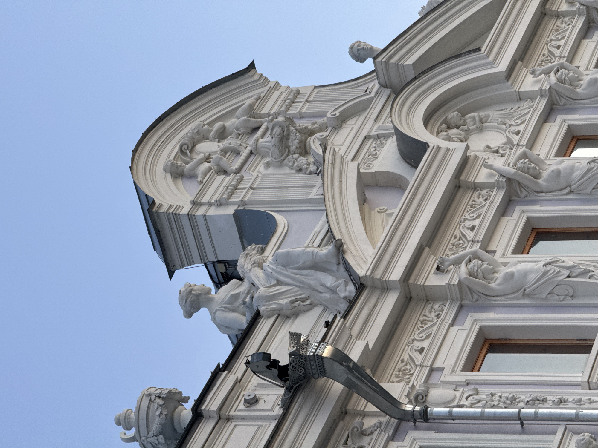 The image shows a detailed view of the facade of the Melnikov House, a historic building located in Nizhny Novgorod, Russia. The architecture features classical elements with ornate sculptural decorations. Prominently visible are white plaster sculptures on the roofline and upper facade. These include several figural sculptures, such as a woman seated with a child, and other female figures supporting architectural elements. Decorative urns and cherubic figures are also present. A metal drainpipe system is attached to the side of the building. The building's facade is painted in light colors, contrasting with the darker roofline. The sky above is a clear, light blue, indicating daytime. The overall impression is one of an elaborate and well-preserved architectural style.