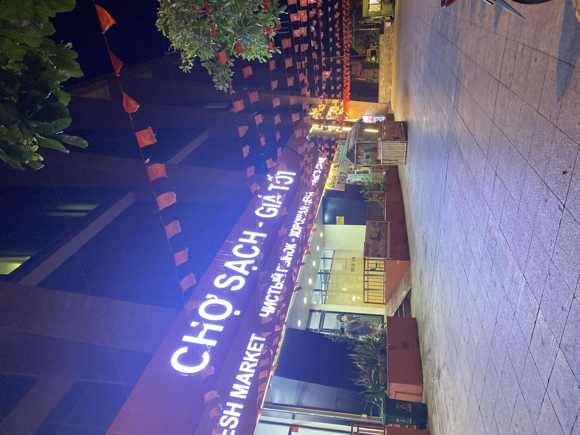The image shows the storefront of a market named "Cho Sach - Gia Tot" (translated as "Clean Market - Good Price"), also translated into Russian as "Чистый Гонок - Хорошо" (which roughly translates to "Clean Chase - Good"). The English text "Fresh Market" is also visible on the sign. There are red flags strung above the entrance. A street is visible next to the storefront with visible sidewalk. The store is in Nha Trang Ward, Vietnam, based on text on the sign.