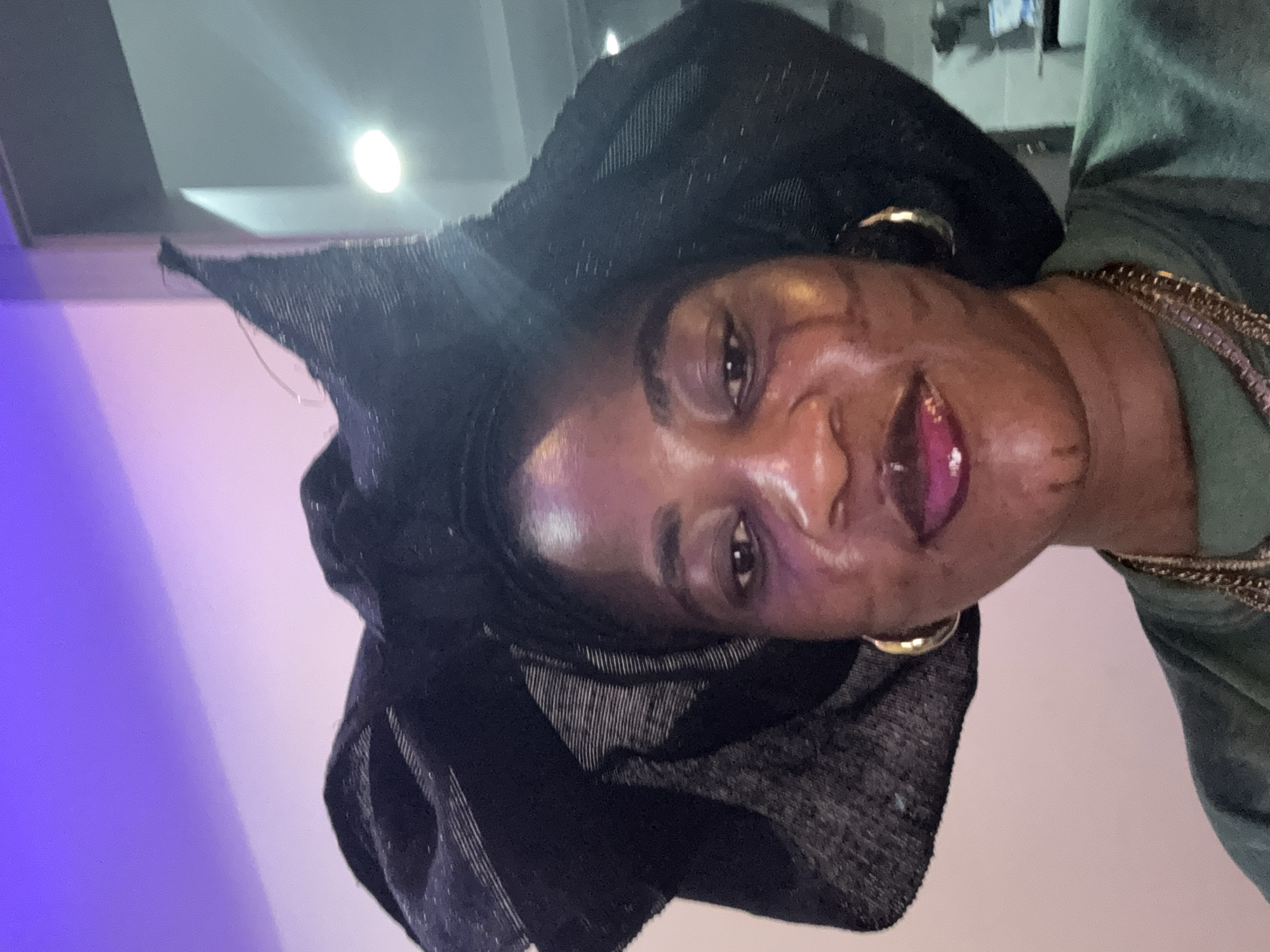 The media file is a vertical, close-up portrait of a Black person wearing a black headscarf and a green top. The person has a gold hoop earring in their visible ear and is wearing a gold chain necklace. Their lips are painted with a vibrant pink lipstick. The lighting suggests an indoor setting, possibly a studio or a well-lit room, with a bright light source visible in the background. The background is predominantly a light pink and purple gradient, possibly from stage lighting, with a sliver of a ceiling and what appears to be a portion of a building or furniture in the upper right. The person's expression is direct and engaging, with a slight smile. The image has a slightly grainy quality. The context provided indicates the location is Ajara, Nigeria. No text is visible in the image.