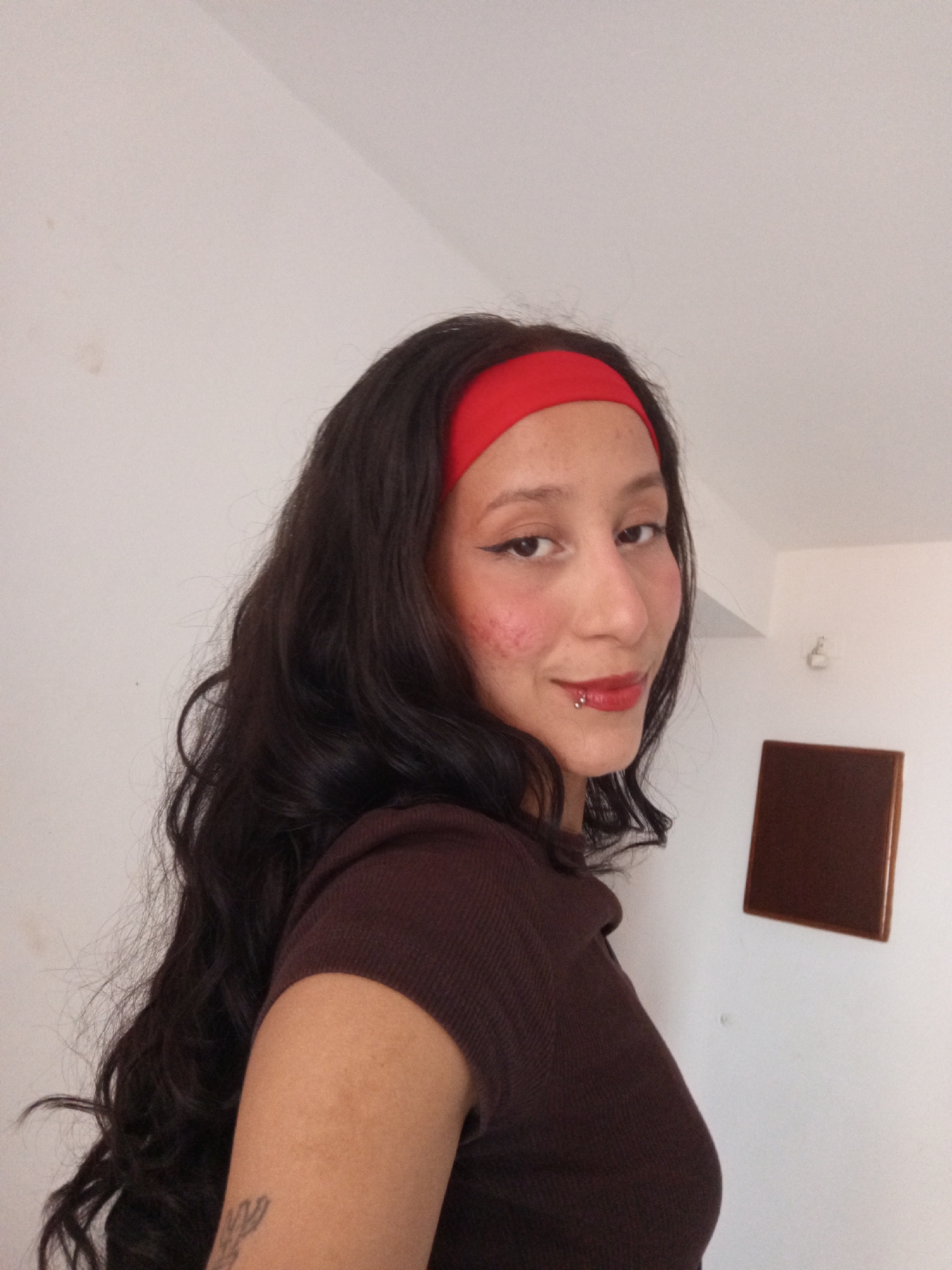 A person with long, dark, wavy hair and a red headband is positioned against a plain white wall. The person is wearing a dark brown short-sleeved shirt. They have a lip piercing and are wearing makeup, including red lipstick and winged eyeliner. Rosy blush is present on their cheeks. A small tattoo is visible on their upper arm. Behind the person, a square, dark brown object hangs on the wall. The location in Porlamar, Venezuela cannot be determined from the image.