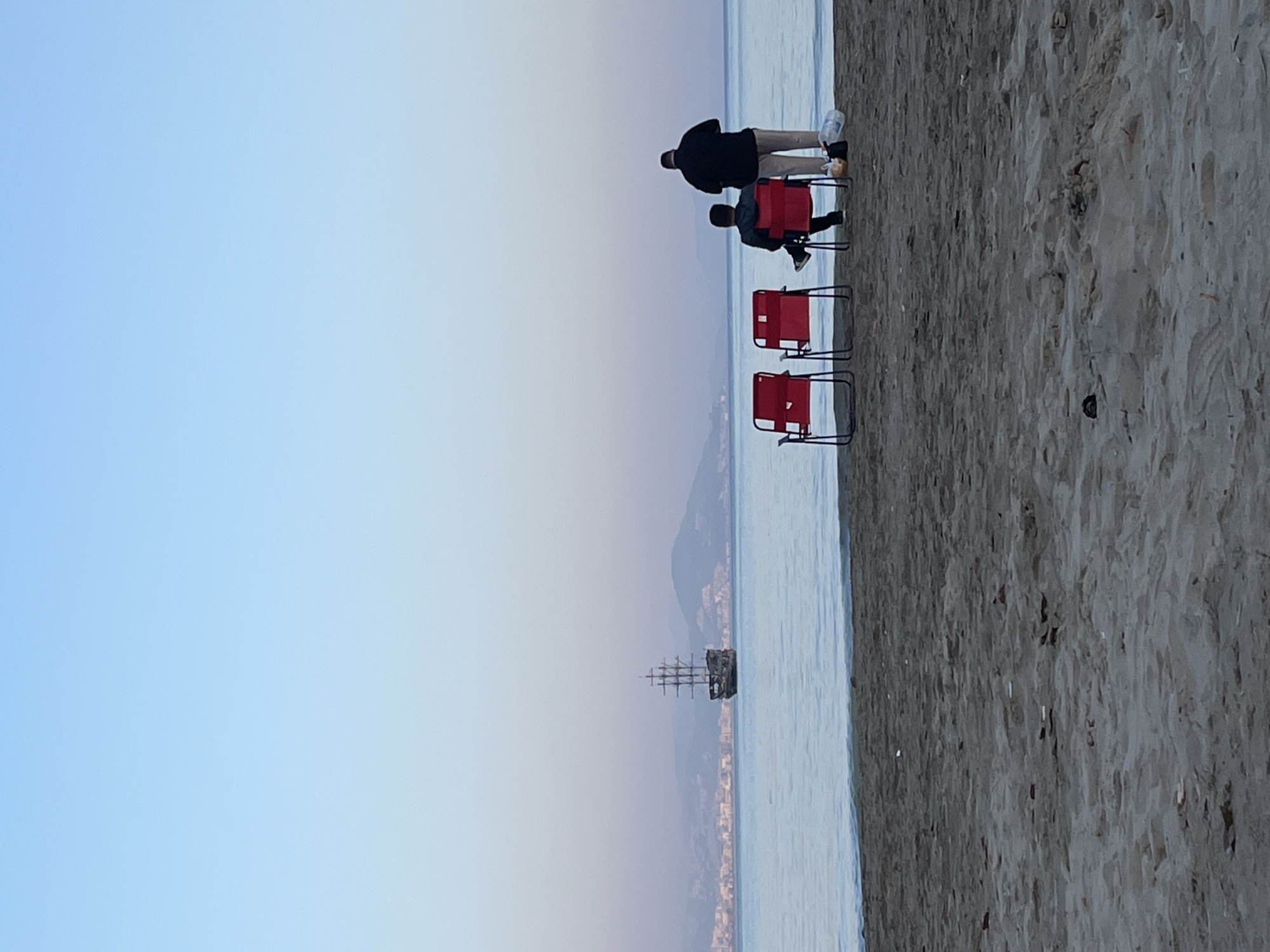 The image captures a tranquil scene on a beach, likely in Alanya, Turkey, given the discernible mountain range in the distance and the presence of a pirate ship replica in the sea. The scene is composed of several key elements:
* **People:** Two individuals are on the beach, one standing and the other sitting on a red folding chair.
* **Objects:** The scene includes three red folding chairs and an empty plastic water bottle. A distant ship with sails is visible on the water.
* **Environment:** The setting is a beach with sand leading to the sea. The sky is light blue with hints of pink near the horizon. There are hills or mountains visible across the sea, with what appear to be buildings along the base of the mountains.
* **Actions/Interactions:** One person is standing, seemingly observing something in the distance, while the other is seated. There are no overt interactions happening, suggesting a serene, observant atmosphere.