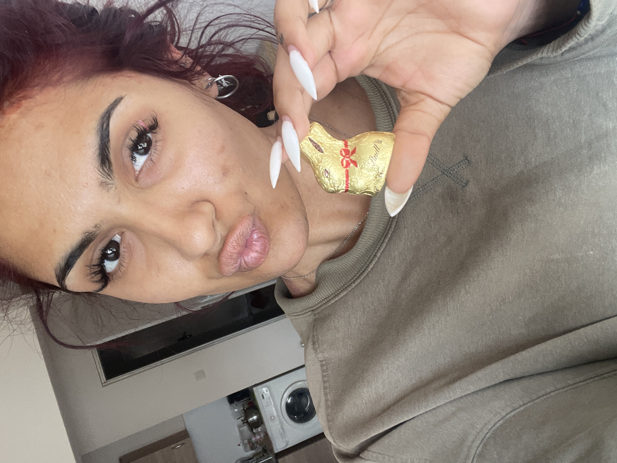 A rotated medium shot depicts a woman holding a gold foil-wrapped chocolate Lindt bunny in her right hand. She has medium skin tone, dark brown hair with red highlights, and dark eyebrows. She is wearing eyelash extensions and has hoop earrings on her left ear. Her fingernails are long, white and pointed. She is wearing a gray-green sweatshirt. Her lips are pursed into a kiss.
In the background, a washing machine is visible in a room with white walls and a light-colored counter. The location is Unknown, Unknown.