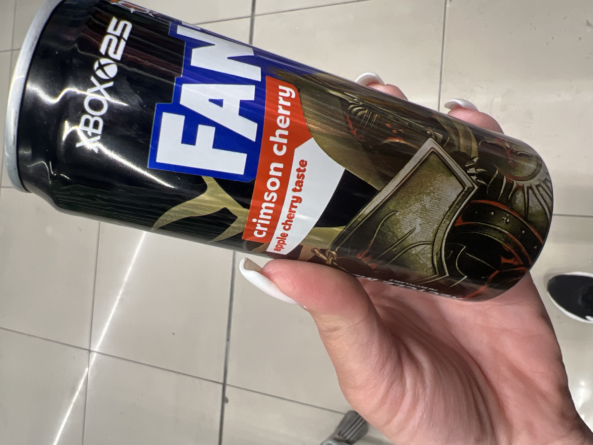 A person with white painted nails is holding an "Xbox 25 Fanatika" Crimson Cherry flavor energy drink can. The can is mostly black with a blue and white "FAN" logo on the front, and an image of an armor-clad character. The background shows a tile floor, and part of a black and white shoe can be seen. The location appears to be inside a store, based on the floor tiles and the presentation of the product. The city is Budva, Montenegro, based on the availability of "Fanatika" brand energy drinks.