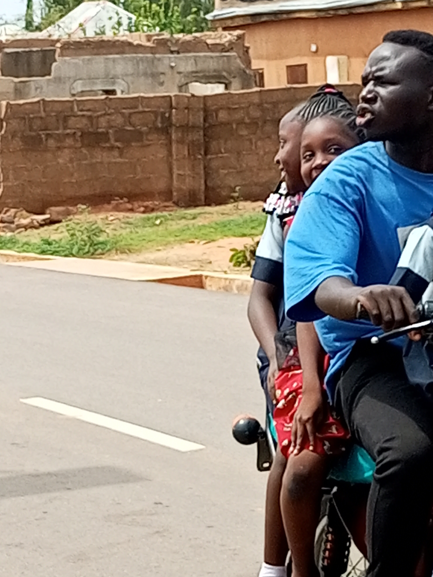 This is a photograph of four people riding on a motorcycle. The person in the front, a man, is wearing a blue t-shirt and black pants, and is driving the motorcycle. Behind him, a girl in a red and white patterned skirt is seated, followed by another child. The fourth person, mostly obscured, appears to be another child in the back. They are riding on a paved road in a street scene in Gombe, Nigeria. In the background, there are partially constructed brick buildings and some green vegetation. The overall impression is that of a typical mode of transportation in an urban environment.