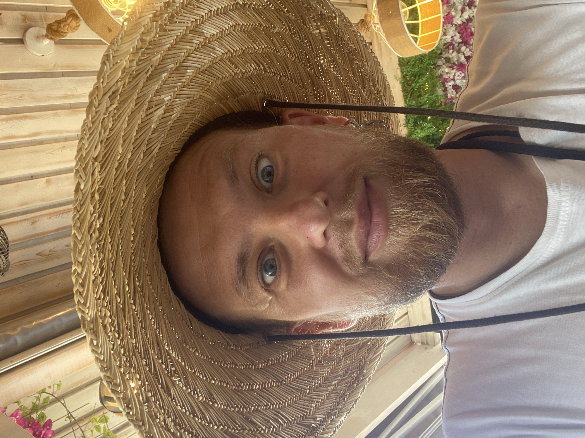In a close-up, low-angle selfie, a fair-skinned adult male with a light brown beard and blue eyes wears a wide-brimmed straw hat that obscures the top of his head and the background. His eyes are wide open, looking directly at the camera. He is wearing a white t-shirt, and black drawstrings from the hat are visible, one on either side of his face. The background appears to be an outdoor setting, possibly a restaurant or cafe, with a wooden slatted ceiling and decorative foliage and flowers. A straw-colored lampshade with a yellow lightbulb is visible in the upper right corner. The overall impression is of a casual, sunny day. The provided information does not specify a precise location within Ia Municipal Unit, Greece, but the setting suggests a tourist or leisure area.