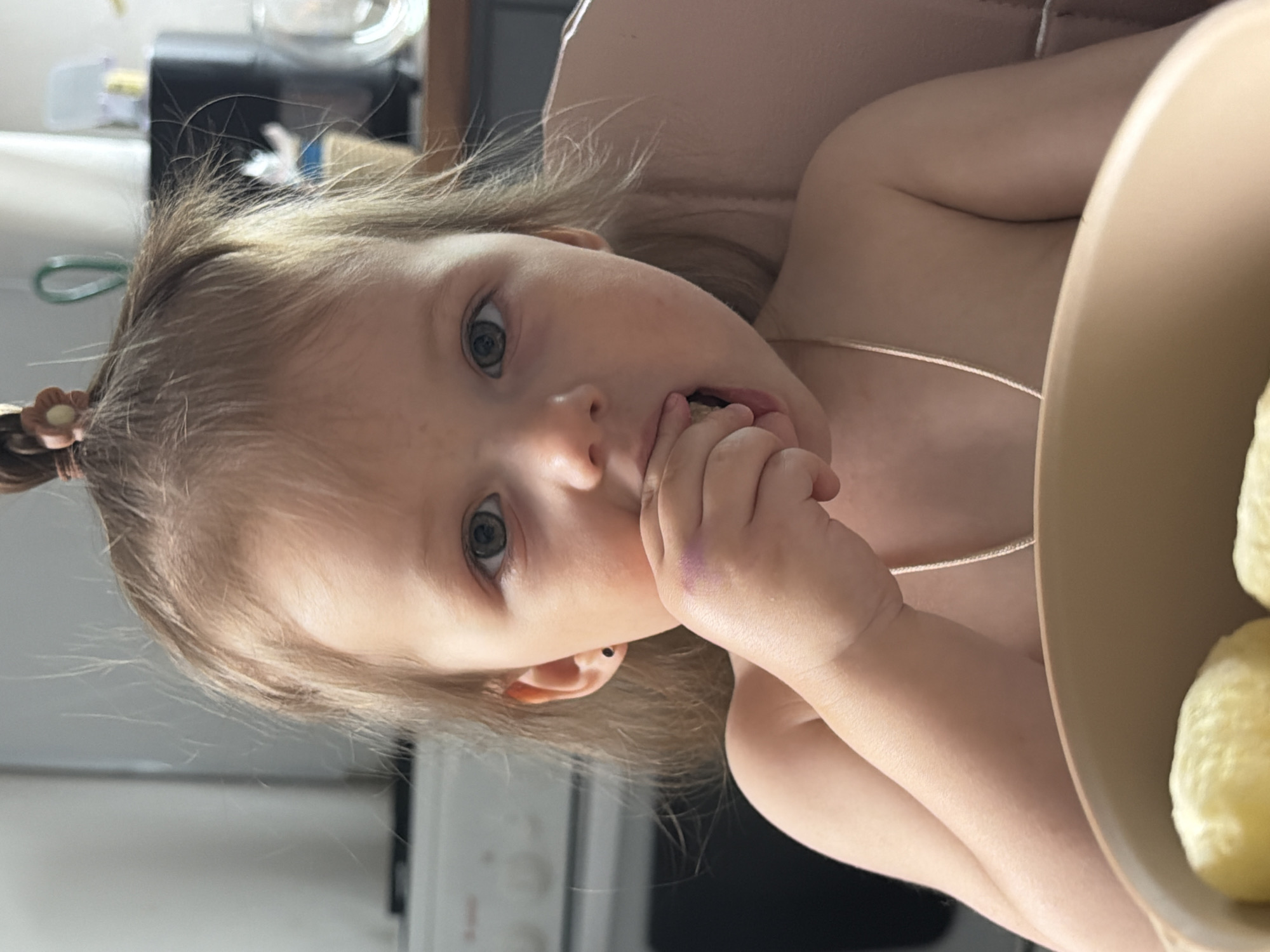 A toddler with fair hair tied in a ponytail, wearing a thin gold necklace, is seated at a table, eating something with their left hand. A purple mark is visible on the child's left hand. A bowl with pale yellow food is present at the right edge of the frame. The background includes a white appliance and other kitchen objects. There is no information in the image that indicates the location is Reshetnikovo, Russia.
