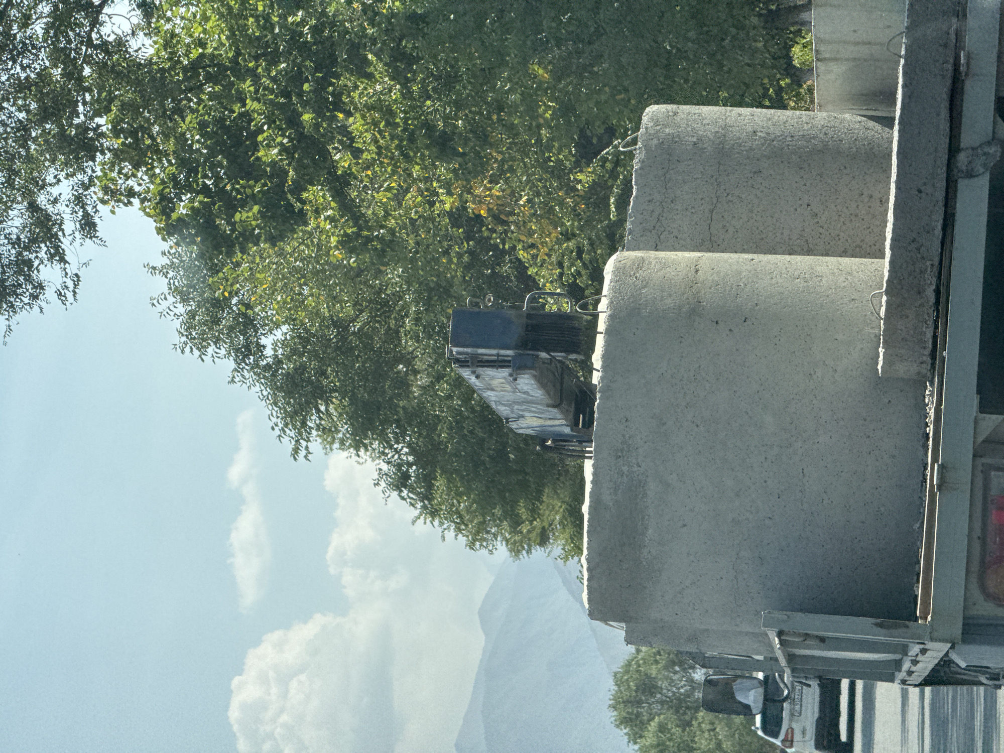 In Tash-Moinok, Kyrgyzstan, a flatbed truck carries several large, cylindrical concrete pipes. The truck is on a paved road, and behind it, another vehicle, a white SUV, is visible. The scene is set against a backdrop of lush green trees and distant mountains under a bright, cloudy sky.