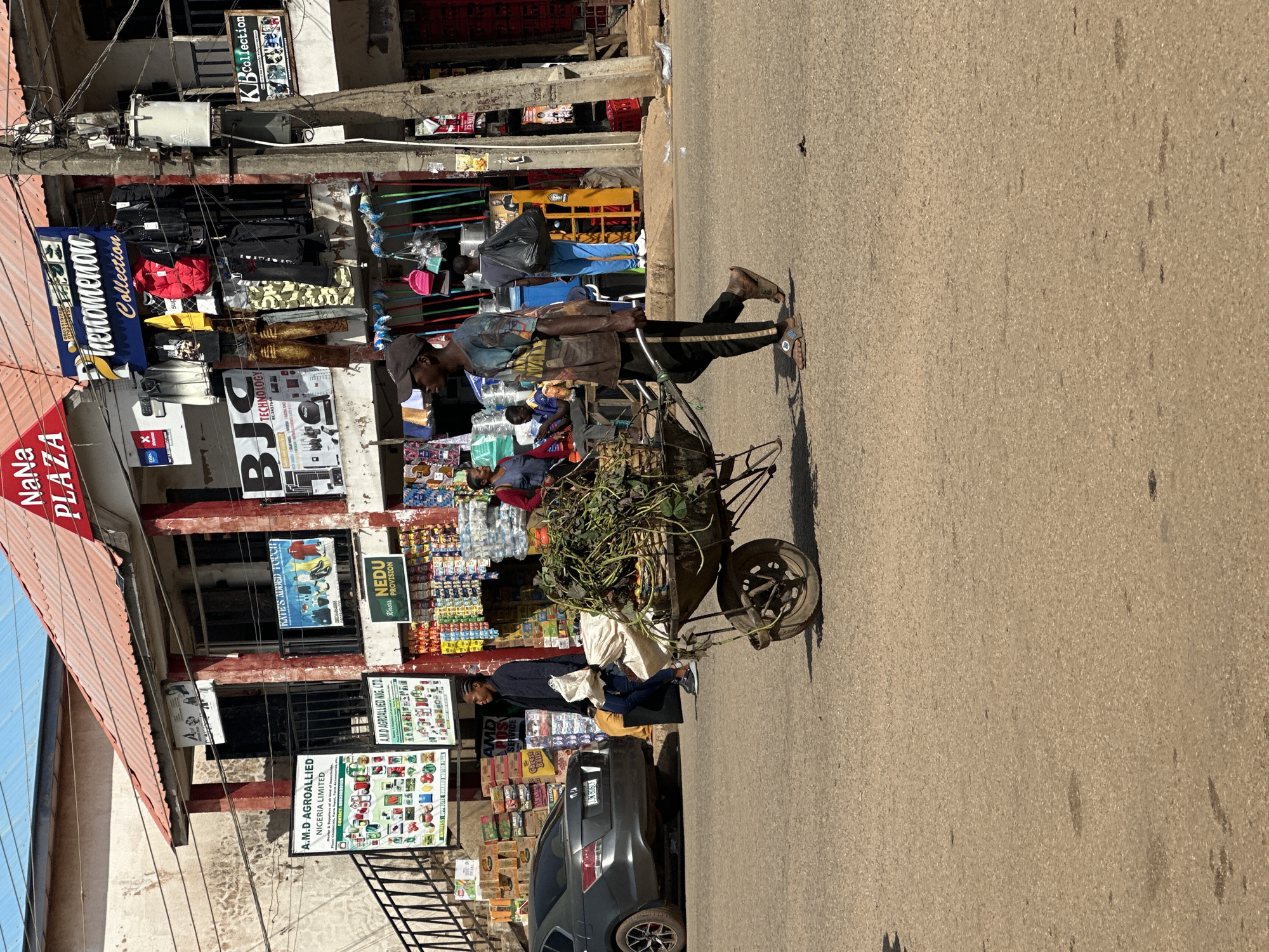 In Bukuru, Nigeria, a man is pushing a wheelbarrow filled with foliage and what appear to be agricultural products down a street. The wheelbarrow is laden with green leafy vegetables, some root vegetables, and other assorted items, possibly produce for sale. The man wears a patterned t-shirt, dark pants, and sandals. He is walking on a paved road, which is slightly worn and shows some dark stains.
To the right of the man, a multi-story building houses various shops. Above the shops, signs read "NaNα PLAZA" and "Phenomenon Collection". Clothing items are displayed outside one of the shops. To the left of the man, a grey sedan is parked on the side of the road. In the background, more shops and buildings are visible, with an array of goods displayed, including packaged products and bottles. The environment suggests a busy commercial area. The sky above is clear and blue.