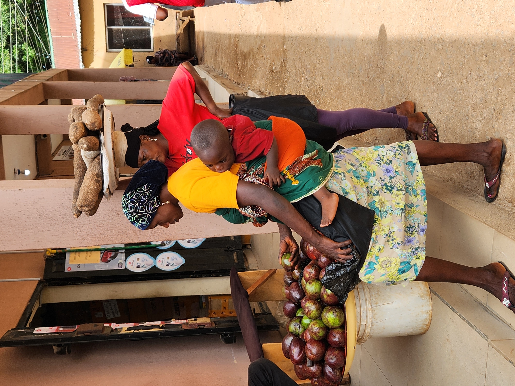 A woman carries a child in the street, with a bucket of avocados to her right. A second woman walks behind. The location is Unknown, Unknown.