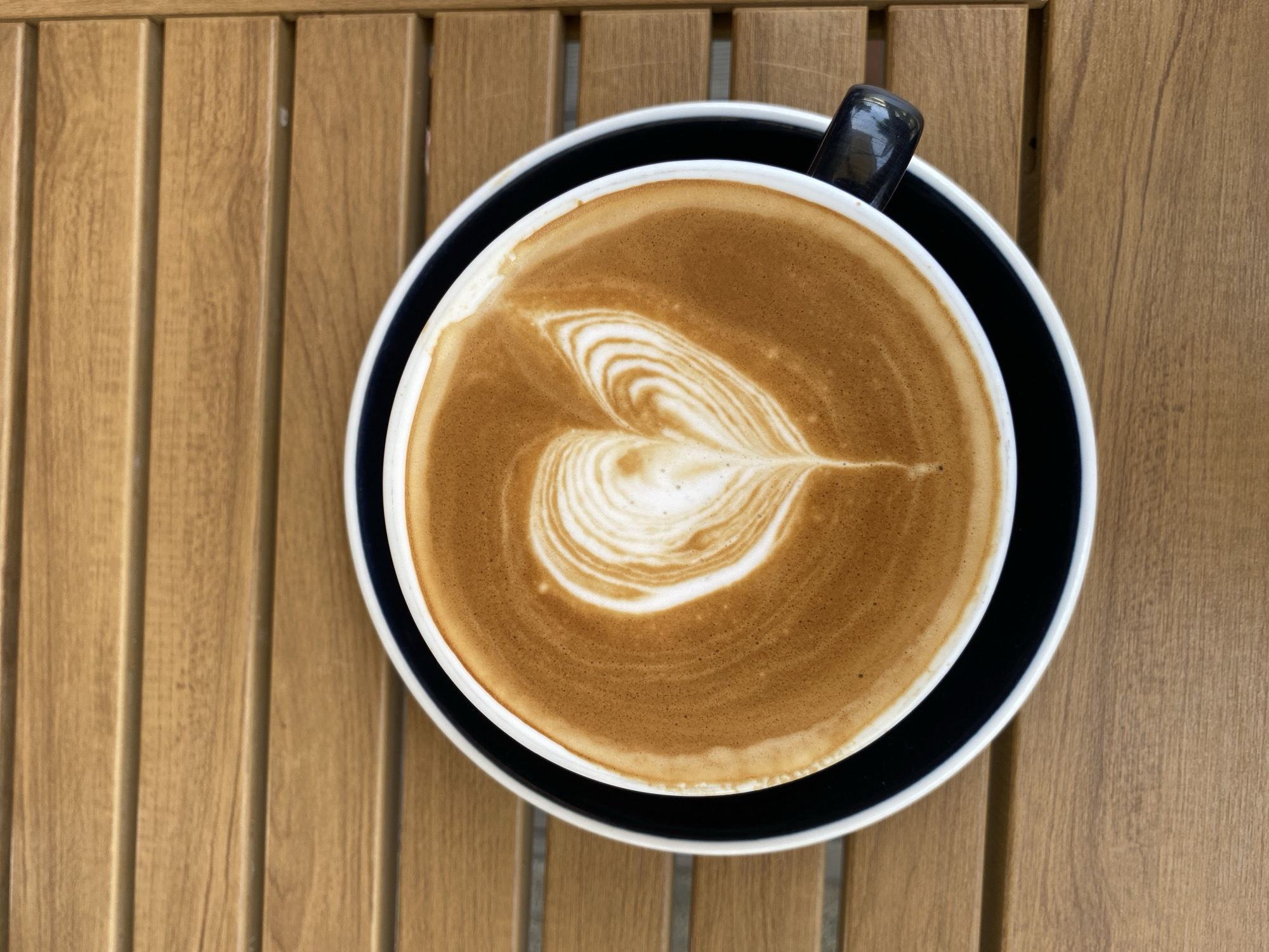 A top-down view shows a cup of coffee with latte art on a wooden table. The coffee is in a white cup, set on a black saucer with a white rim. The latte art features a heart-shaped design in the center. A black handle is visible on the right side of the cup. The table is made of light brown wooden slats arranged vertically. The scene appears to be outdoors, with natural lighting. The location is Choeng Thale, Thailand, based on external information.