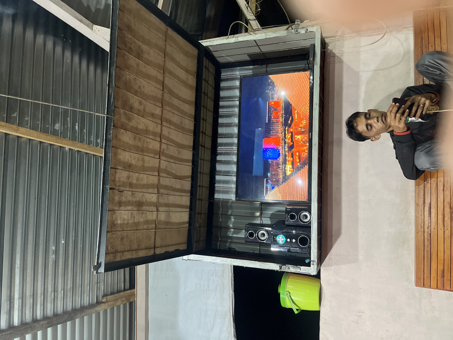 An outdoor TV setup features a large screen displaying a cityscape, along with a speaker system. The setup is enclosed within a metal and wood structure. A man is seated nearby, looking at his phone. A green bucket is visible below the TV enclosure. The surrounding environment appears to be a partially enclosed or outdoor space with metal siding.