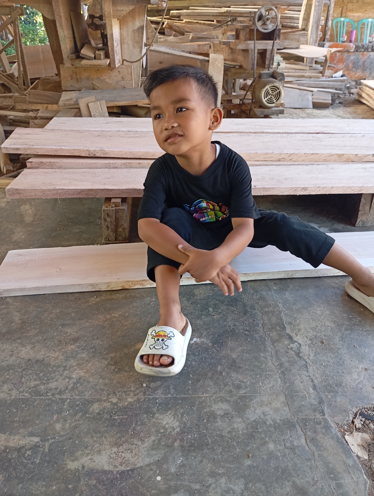 A young boy, appearing to be of Southeast Asian descent, is seated on a stack of lumber. He is wearing a black t-shirt with a colorful graphic and black pants, along with white slip-on sandals. His hands are clasped in his lap. The setting appears to be a workshop or storage area, with piles of raw lumber visible in the background, along with some woodworking machinery. The floor is concrete. The overall impression is a candid shot taken in an industrial or rural setting. The location specified, Ketepung, Indonesia, is noted.