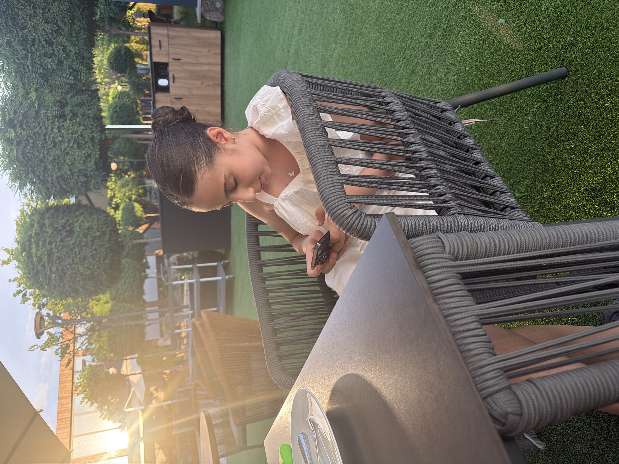 A young girl with dark hair pulled into a bun sits in a gray rope-wrapped chair at a dark gray table. She is wearing a white dress and looking down at her mobile phone, which she holds in her hands. The scene appears to be outdoors, possibly at a restaurant or cafe, with a green artificial turf ground. In the background, there are trees, outdoor furniture, and buildings, suggesting an urban or suburban setting. The sun is low in the sky, casting a warm glow and lens flares. The image is taken in the city of Ashtarak, Armenia.