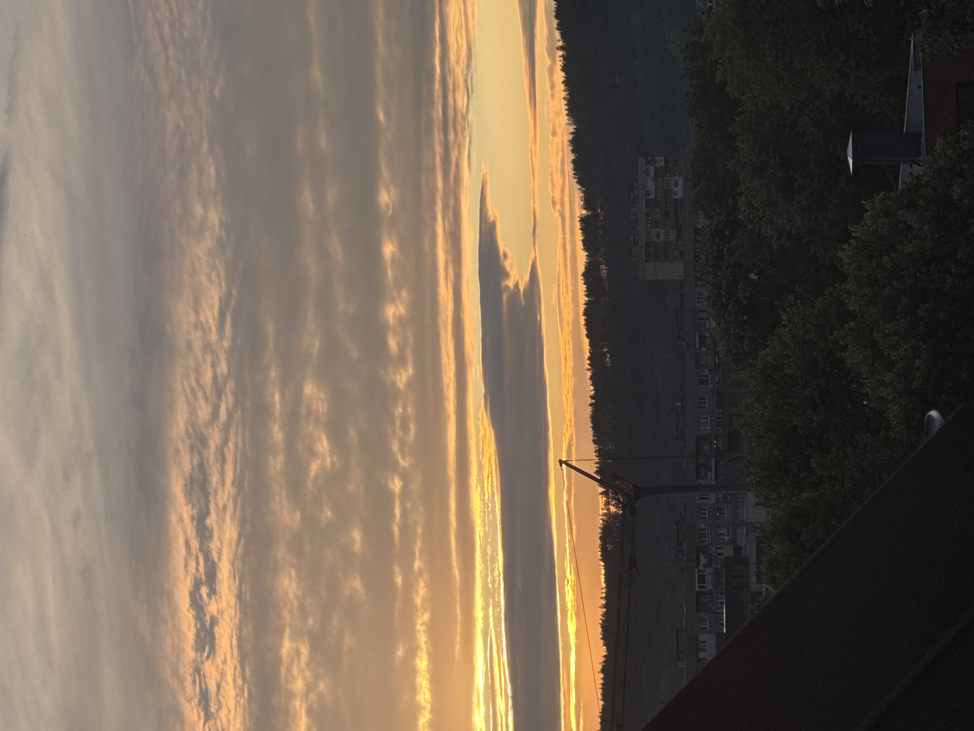 This is a vertical, medium-shot photograph taken at sunset in Pernik, Bulgaria, capturing a silhouetted urban landscape against a dramatic sky. The sky is filled with layered, wispy clouds illuminated by the setting sun, casting a warm orange and yellow glow across the upper portion of the image. Below the sky, a dark, undulating line of trees forms the horizon. In front of the trees, a cluster of low-rise buildings and a prominent construction crane are visible in silhouette. The crane, a large, industrial structure, stands out with its long boom angled upwards. The foreground is dominated by the dark, angled roof of a building, obscuring the lower part of the scene. The overall impression is one of a city at dusk, with the natural beauty of the sunset contrasting with the man-made elements of the urban environment.