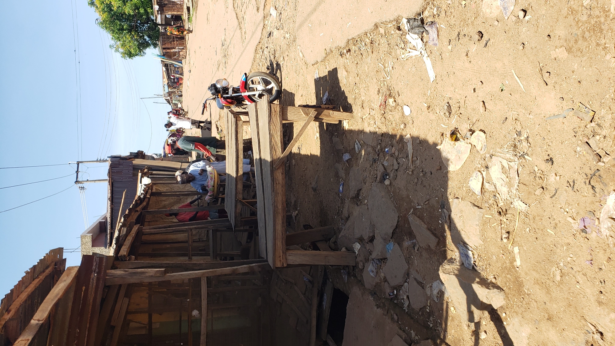 This image captures a street scene in Sabon Tasha, Nigeria, under a clear blue sky. The foreground consists of an unpaved, sandy-brown dirt ground, extensively littered with various pieces of refuse, including plastic bags, paper, and debris, alongside scattered rocks and broken concrete. Two rudimentary wooden market tables, with plank tops and simple leg supports, are positioned prominently in the mid-left, casting sharp shadows towards the viewer.
To the left, a row of makeshift market stalls or shops with wooden frames and corrugated metal roofing stretches into the distance. One stall features a mesh wall. In the midground, several individuals are present. A man dressed in light-colored traditional attire, possibly white or light blue, stands behind one of the wooden tables, holding a large, round tray containing items for sale. Adjacent to him, a man in a green long-sleeved shirt and a red cap stands, looking towards the man with the tray. Further down the unpaved road, additional people are walking and standing.
A red and blue motorcycle with a chrome headlight is parked on the right side of the dirt road, facing away from the viewer. The road surface is uneven, dry, and dusty, with visible tire tracks. Utility poles with multiple overhead power lines traverse the upper portion of the image, extending along the street. In the background, more structures, appearing to be simple buildings and stalls, line both sides of the road, which curves slightly to the right as it recedes. A green-leafed tree is visible on the far right side of the background. The strong sunlight creates distinct, dark shadows throughout the scene, suggesting a time of day with high sun exposure.